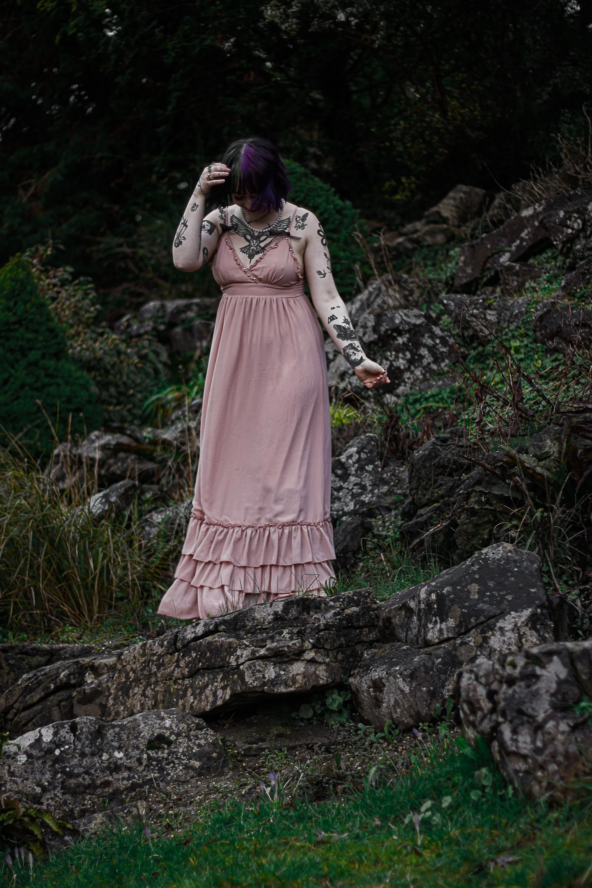 A young woman with purple and black hair, tattoos on her arms and chest, wearing a long pink dress, standing on rocks outdoors surrounded by green foliage.