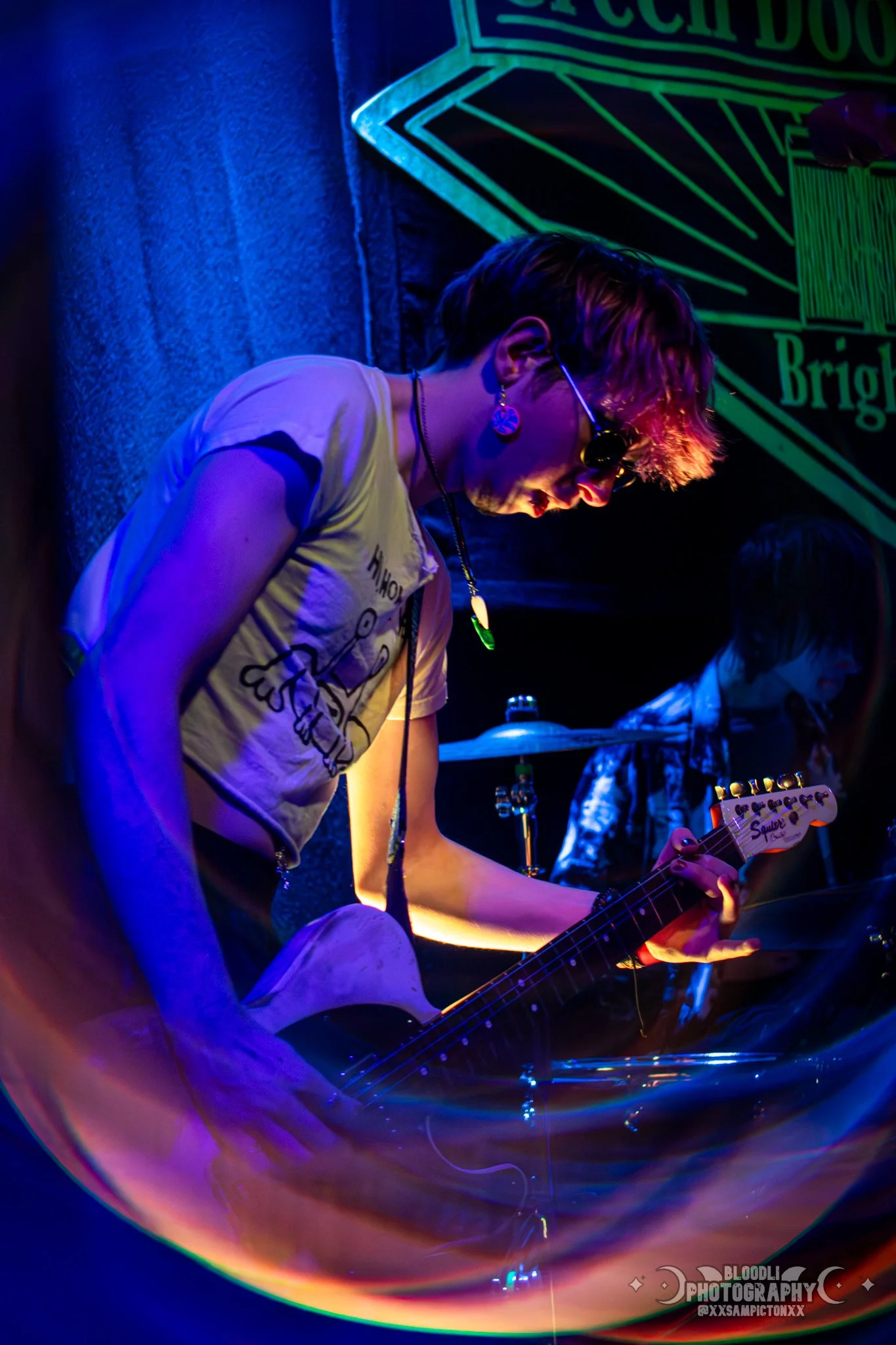 A person playing an electric guitar on stage, illuminated by colorful blue and purple lights, with another musician in the background and a neon sign partially visible behind them.