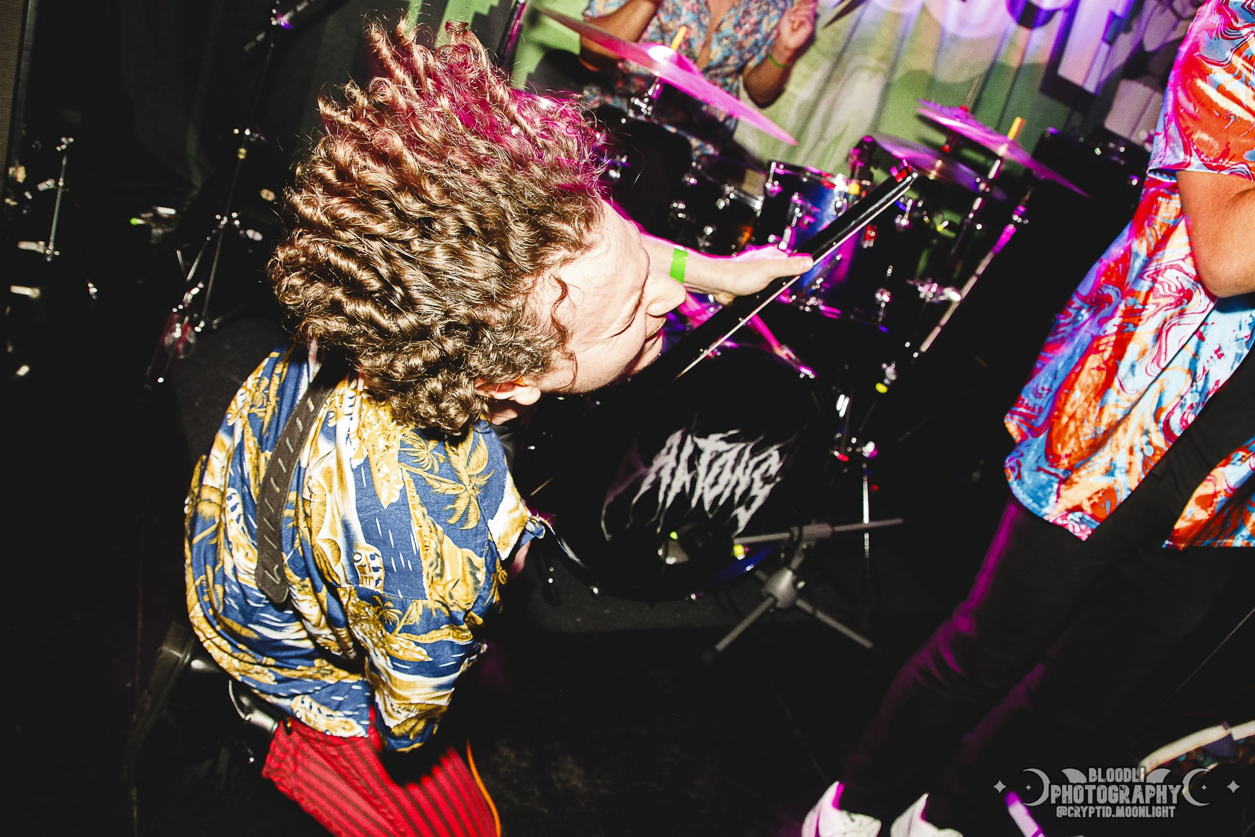 A person with curly hair and a patterned shirt is playing a violin on stage during a live music performance, with a drum set in the background.