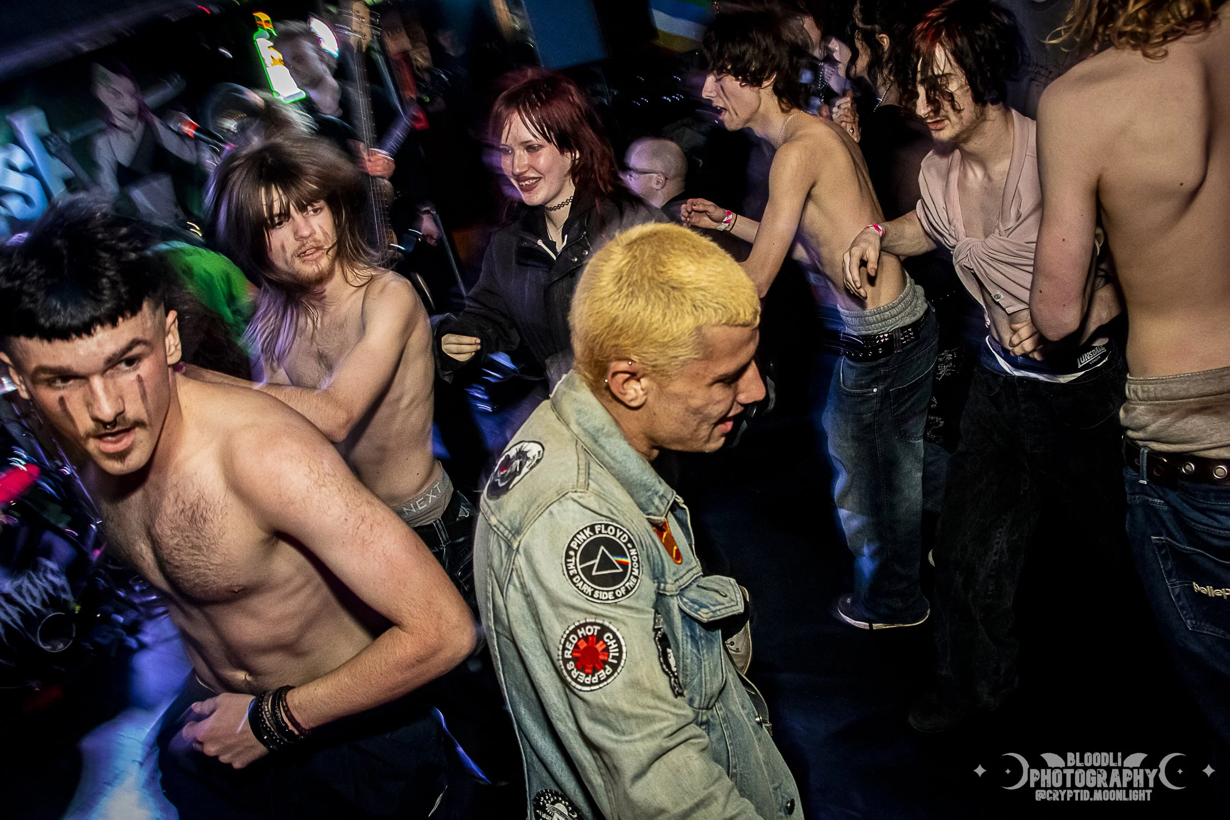 Group of young people dancing in a nightclub, some shirtless, with colorful lighting and neon signs in the background.