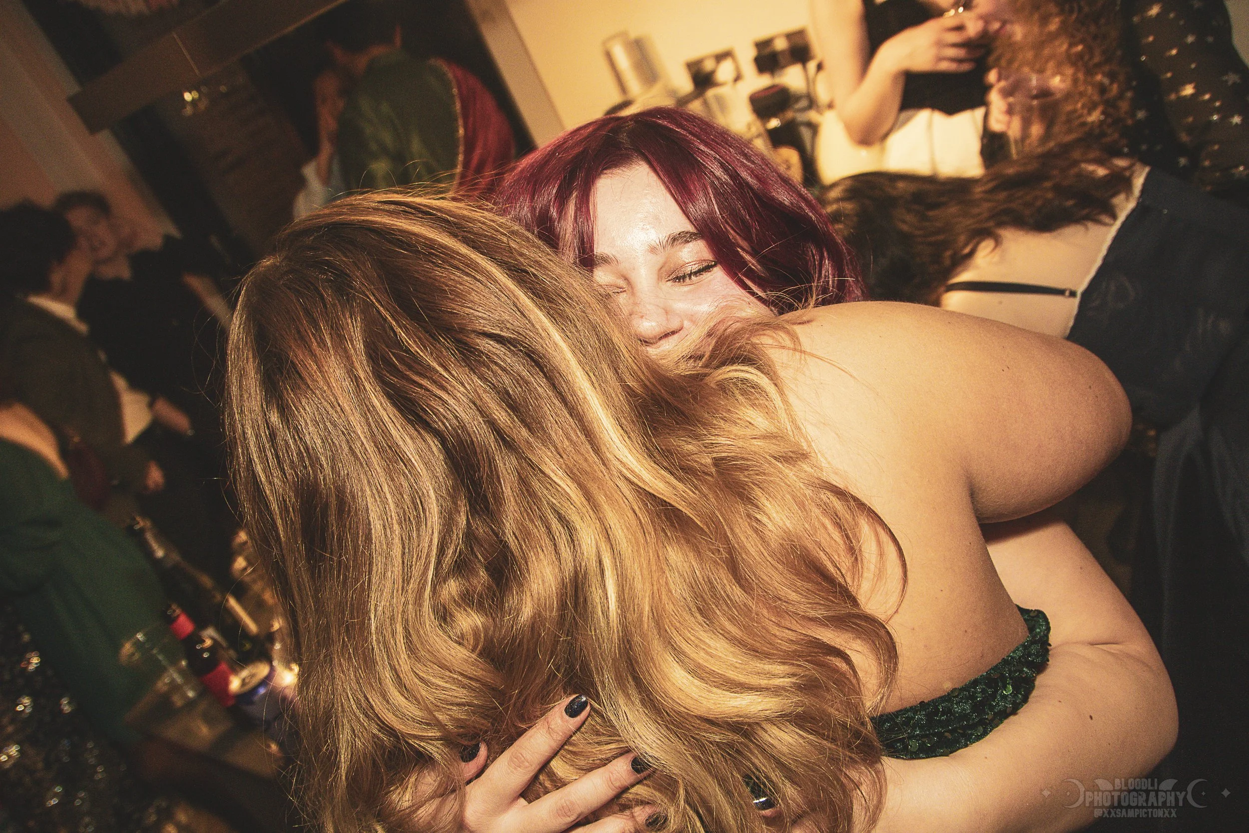 Two women hugging at a social gathering, smiling happily. One woman has long, wavy blonde hair, and the other has short, curly red hair. The background shows other people talking and socializing in a warmly lit room.