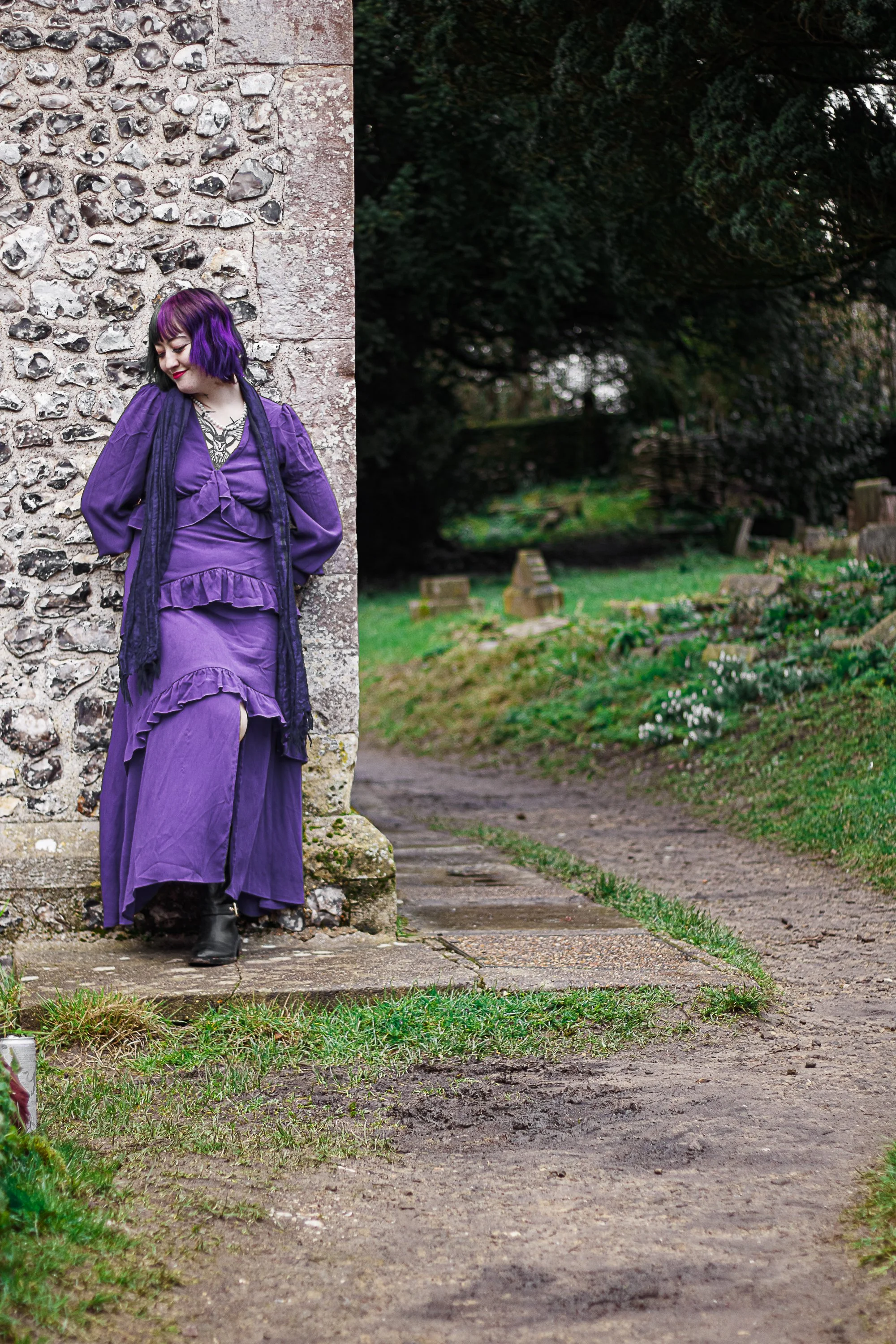 A woman with purple hair and a purple dress standing by a stone wall in a garden.