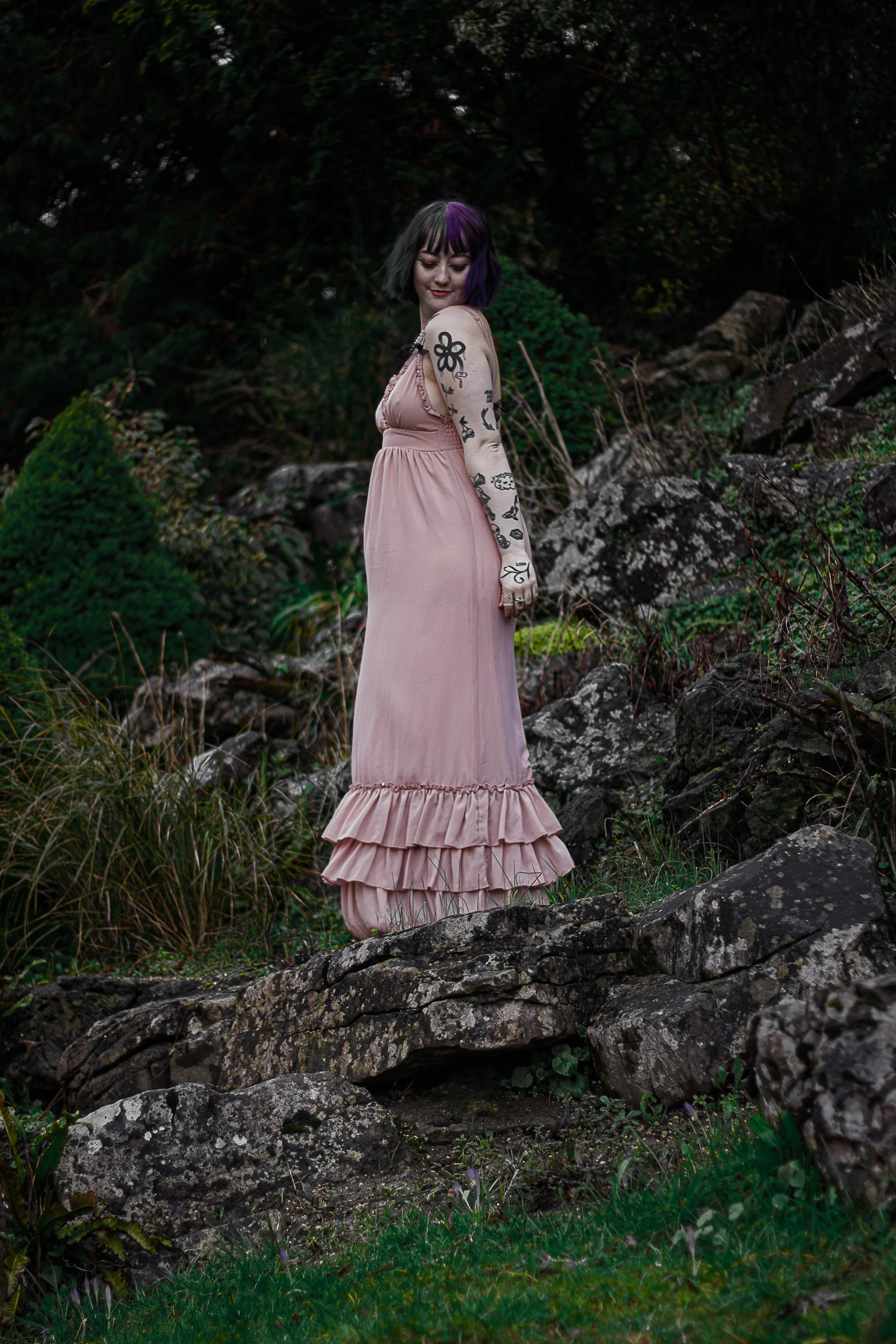 A woman with short black hair and purple highlights, wearing a pink dress, standing on rocks in a natural outdoor setting with greenery and rocks in the background.