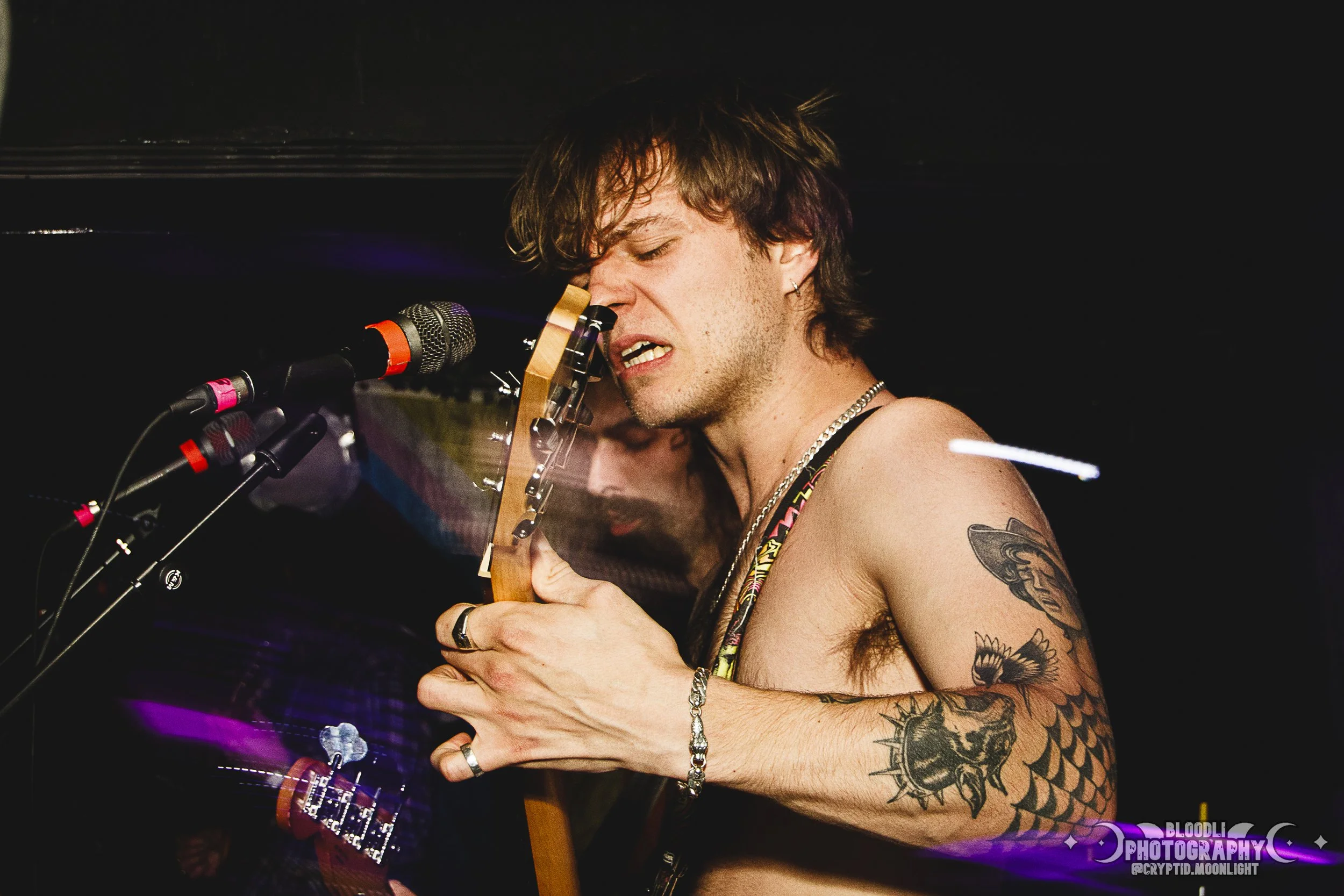 A shirtless male musician with tattoos playing an electric guitar with multiple microphones in front of him.