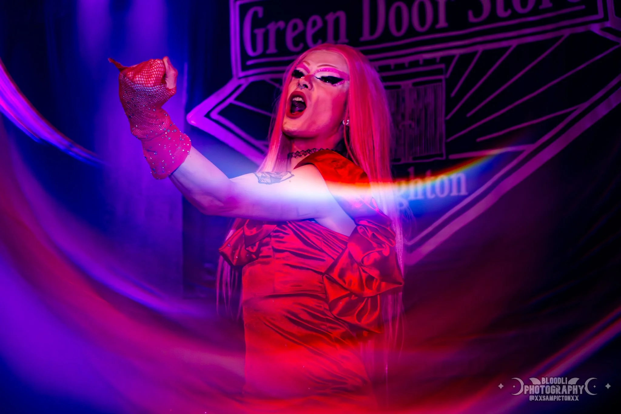 A person with bright pink hair, wearing a red dress and long pink gloves, performing on stage with a purple and pink lighting backdrop, the words 'Green Door Store' visible in the background.