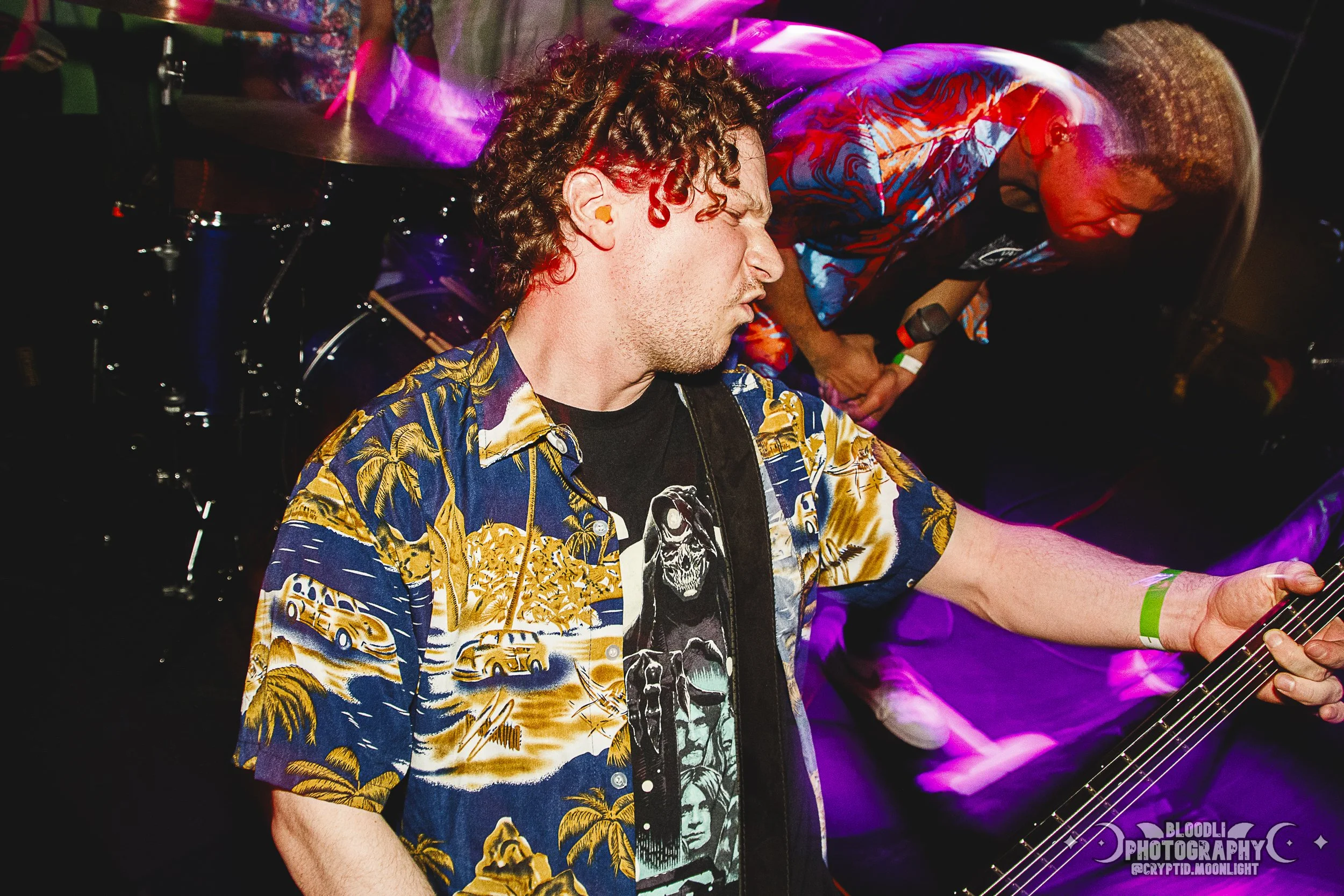 A man with curly hair wearing a colorful Hawaiian shirt is playing an electric guitar, while another person with a shaved head and wearing a tie-dye shirt is in the background, both performing on stage with purple lighting.