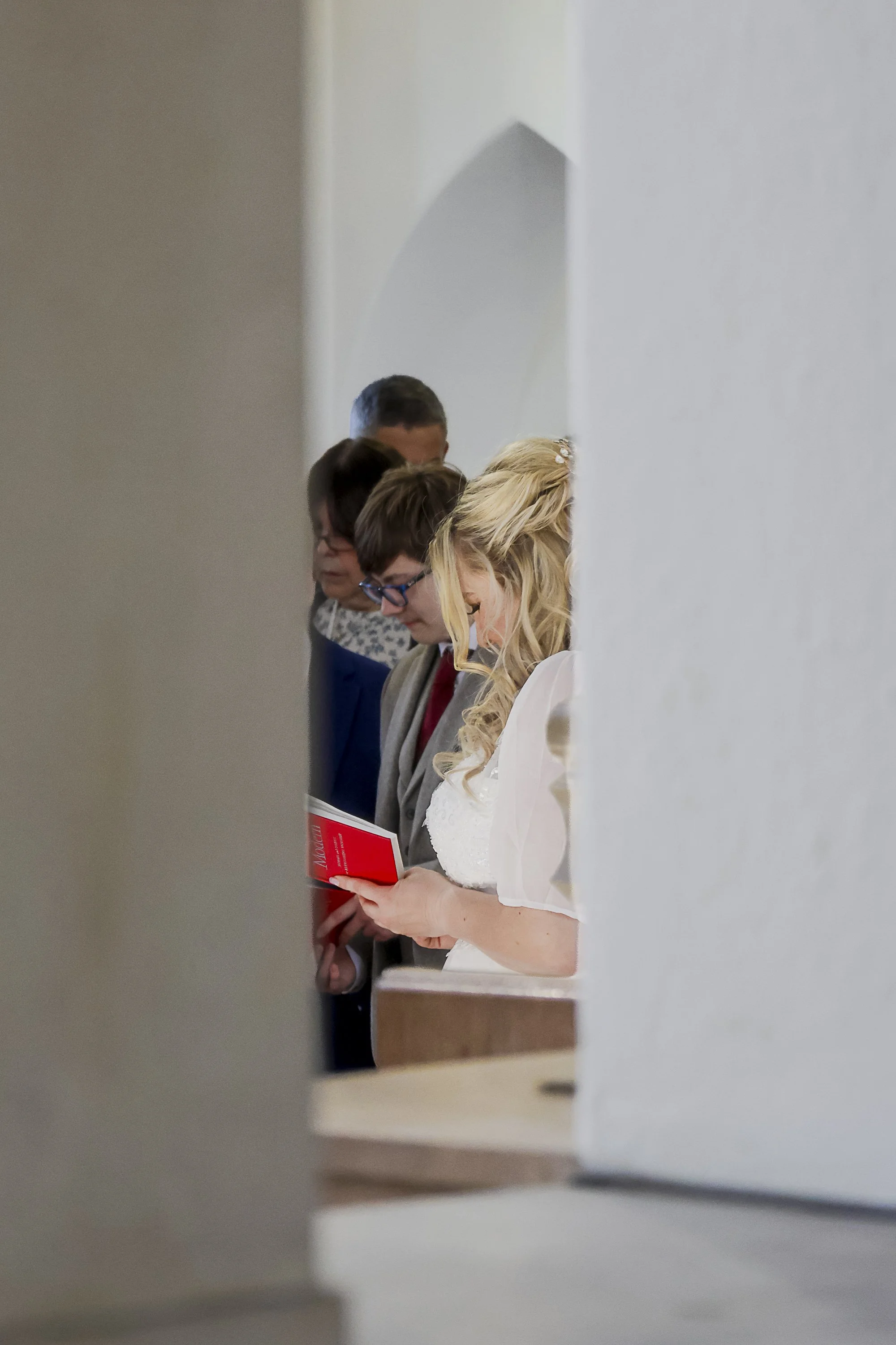 A group of five people, dressed semi-formally, standing in a line, looking down and reading from books or papers, with a plain white wall behind them.
