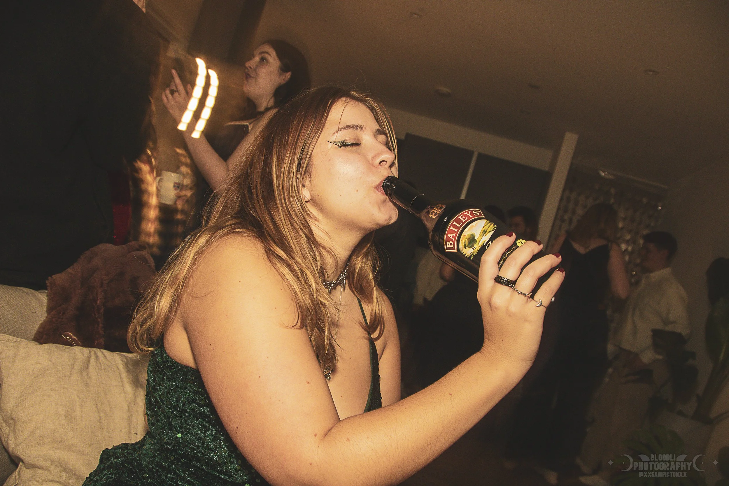 A young woman with long hair, wearing a dark green dress with sequins and jewelry, is drinking from a bottle of Baileys Irish Cream at a party or social gathering. She has her eyes closed and appears to be enjoying herself. In the background, there a