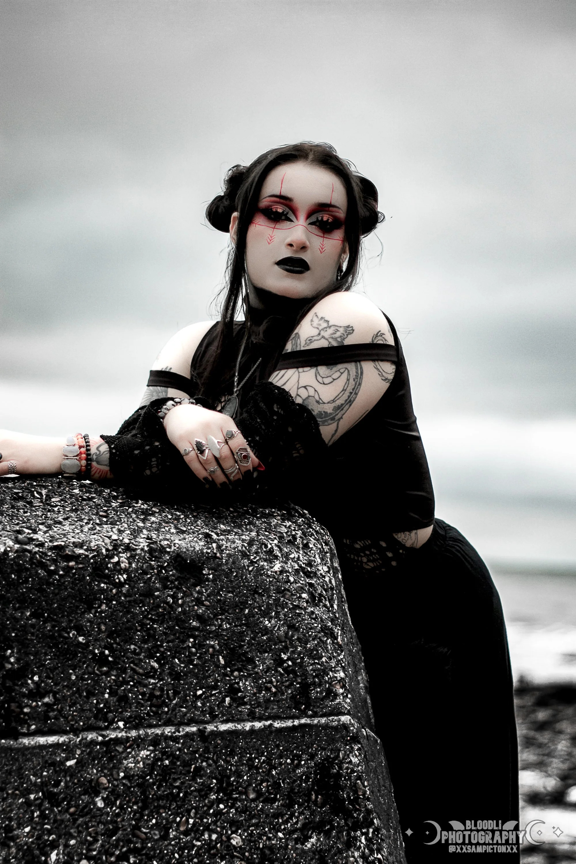 A woman with dark makeup and tattoos leaning on a large textured rock outdoors under a cloudy sky.