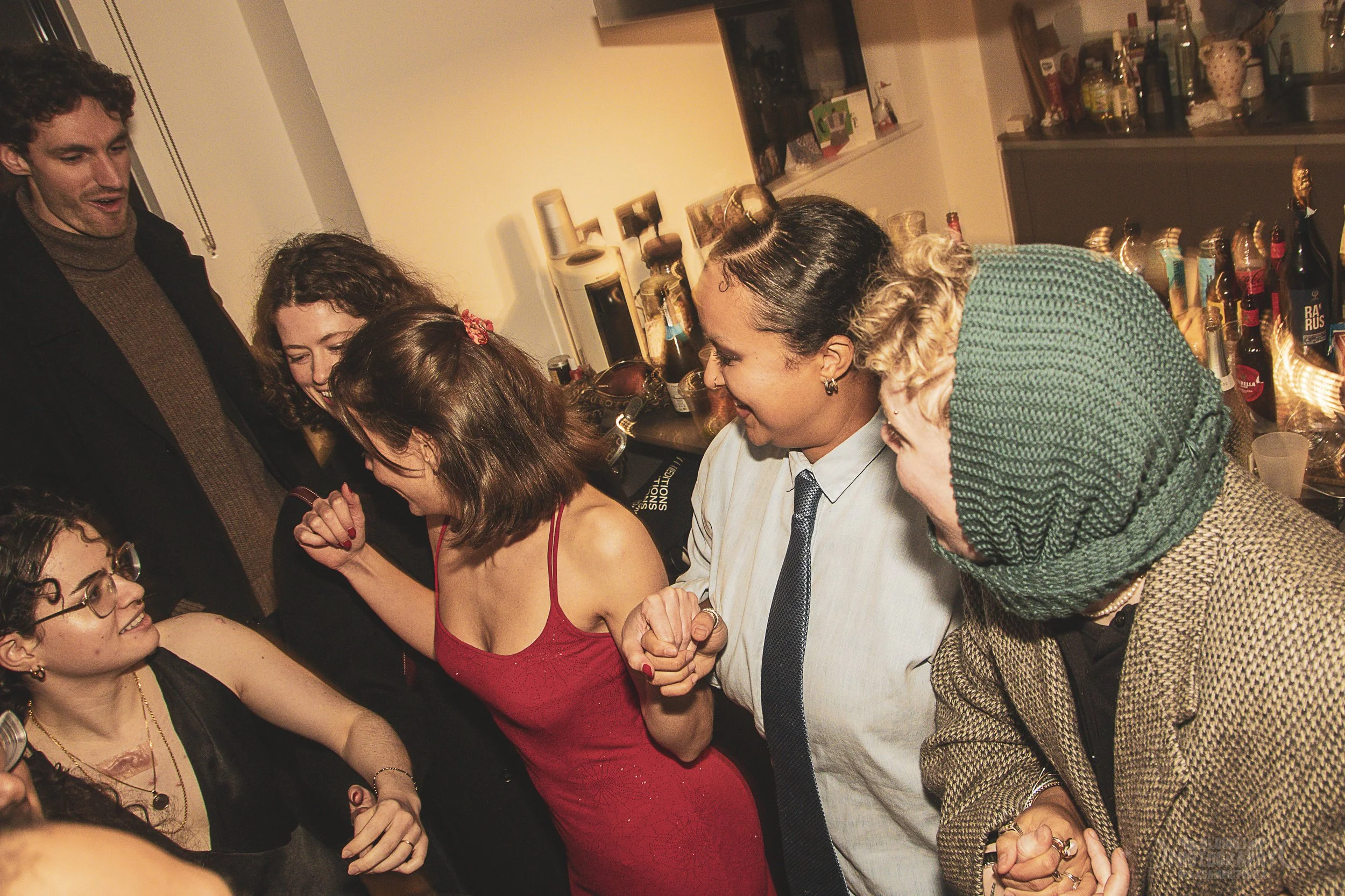 A group of people at a party are holding hands and smiling, with some looking at each other and engaging in playful interactions. The setting appears to be a kitchen or bar with bottles and glasses in the background.