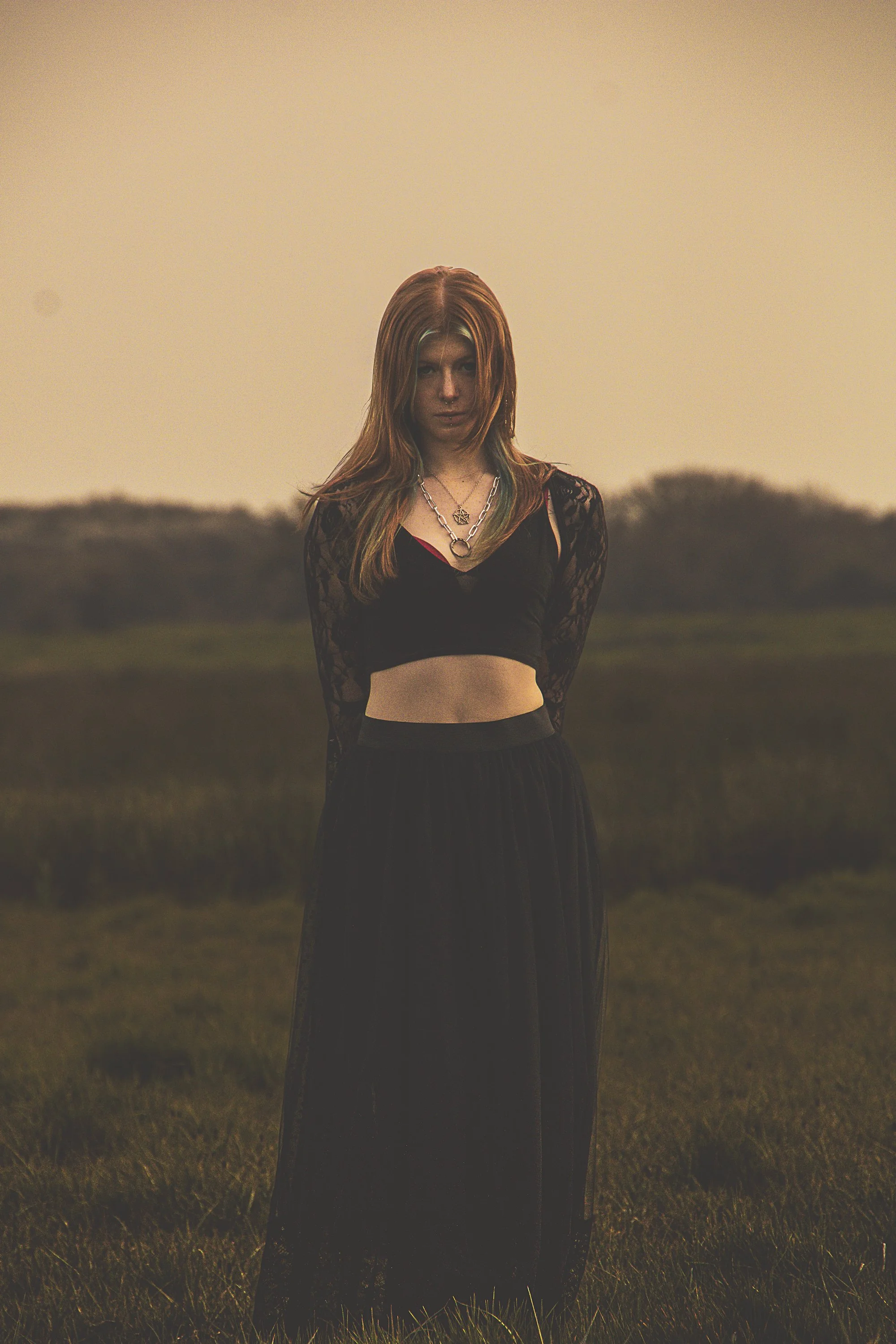 A woman with red hair wearing a black crop top with lace long sleeves, a long black skirt, and layered necklaces, standing outdoors in a grassy field during dusk.