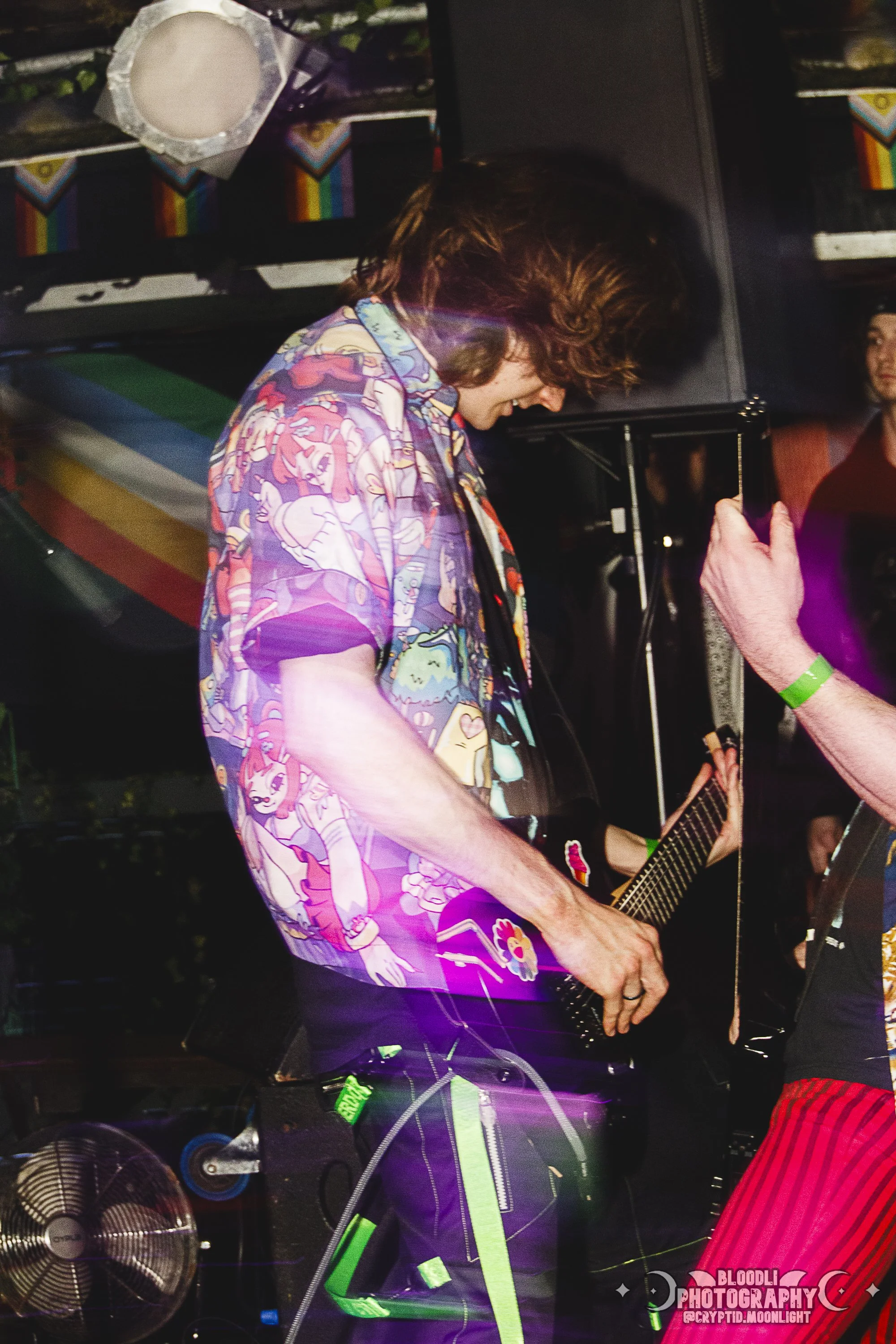 A man playing an electric guitar at a party or concert, wearing a colorful, cartoon-themed shirt, with rainbow flags in the background and purple light effects.
