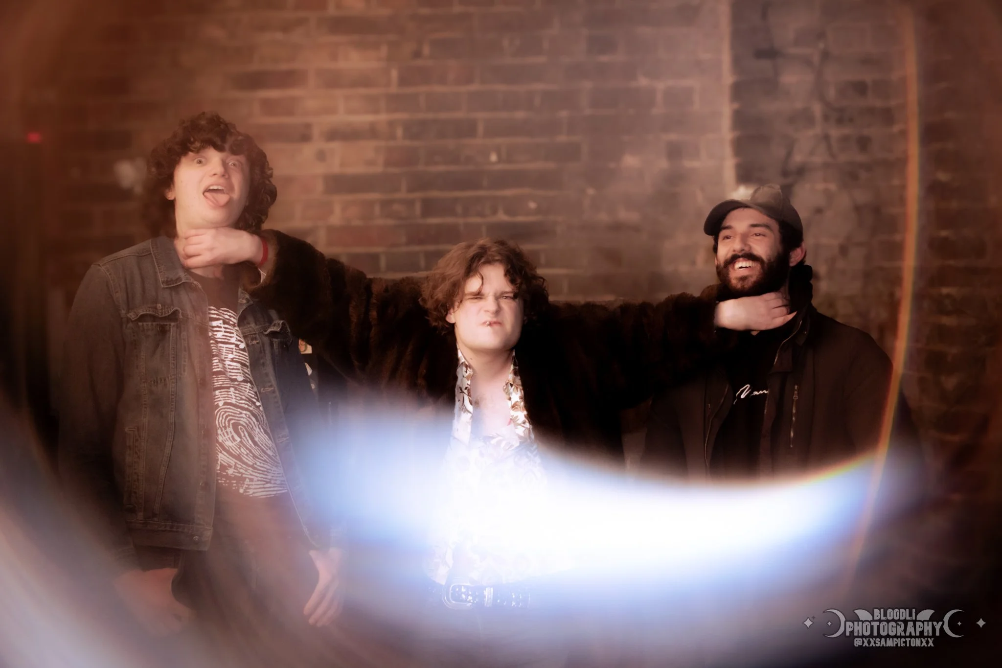 Three men with curly hair and beard posing in front of a brick wall, with the man on the left sticking out his tongue, the man in the middle making a grimace, and the man on the right smiling.