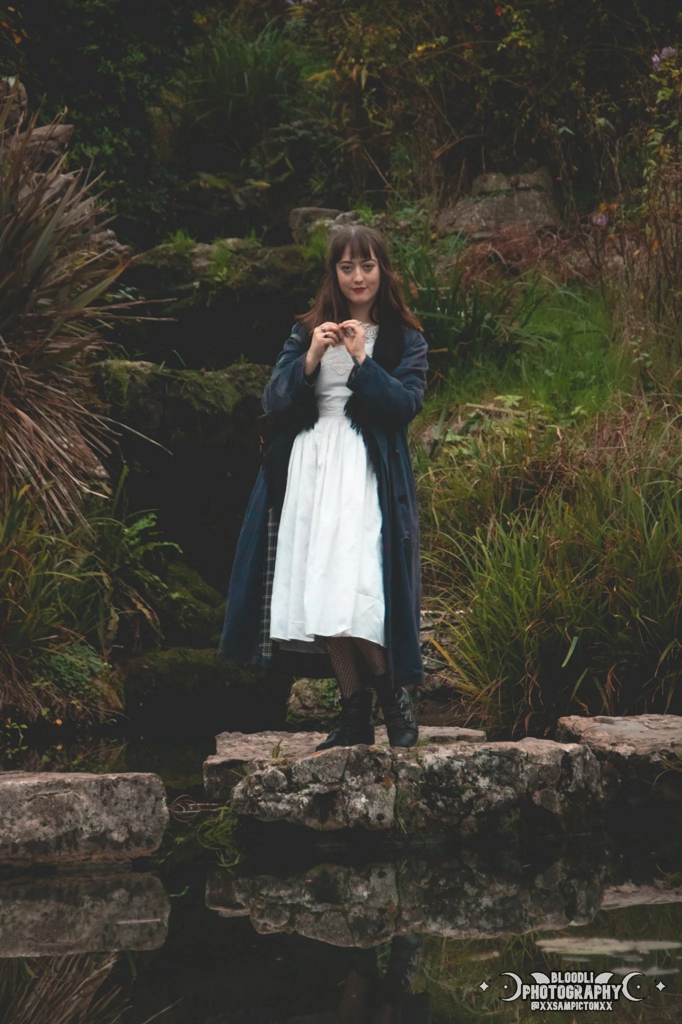 A young woman stands on a large rock in a natural outdoor setting with lush greenery and moss-covered rocks in the background. She is wearing a vintage-style white dress, black fishnet stockings, black boots, and a dark blue coat, with her hands clas