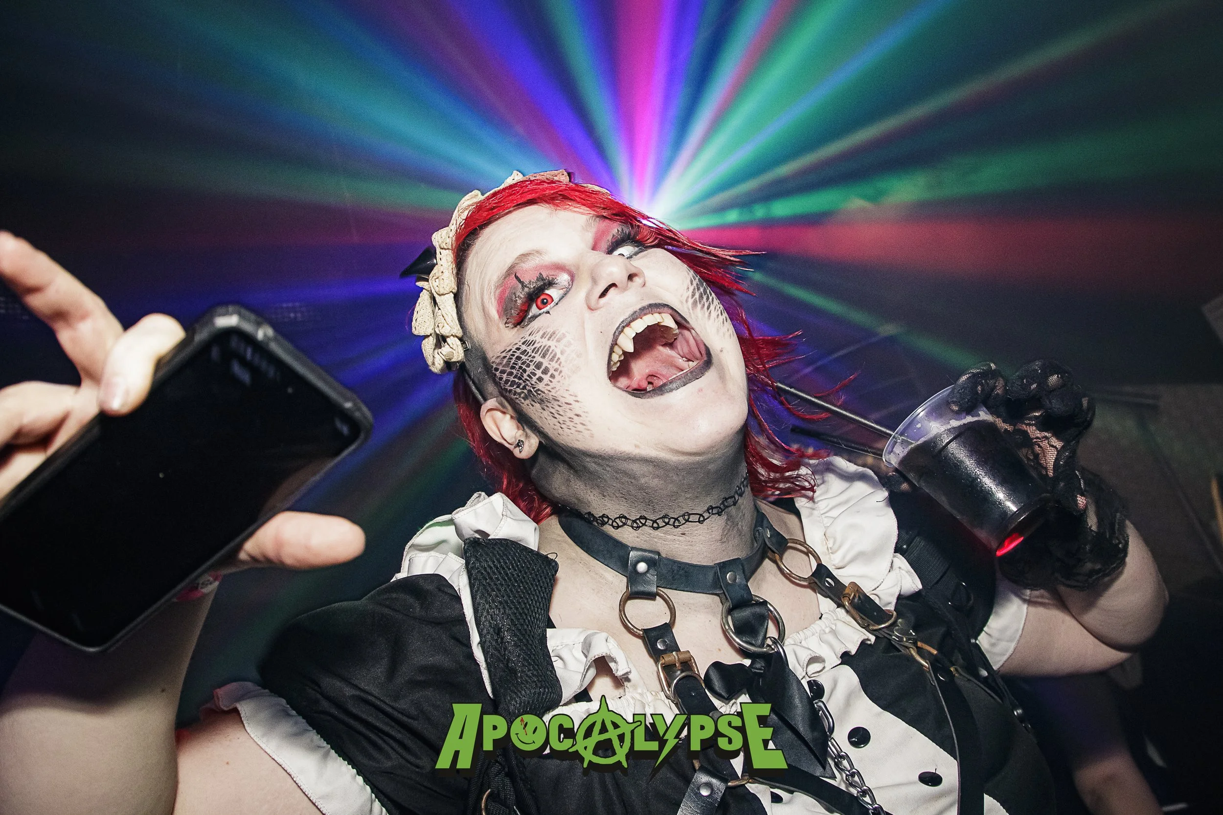 Person with red hair wearing gothic makeup, a choker, and a costume, holding a drink in one hand and taking a selfie at a party with colorful laser lights in the background.