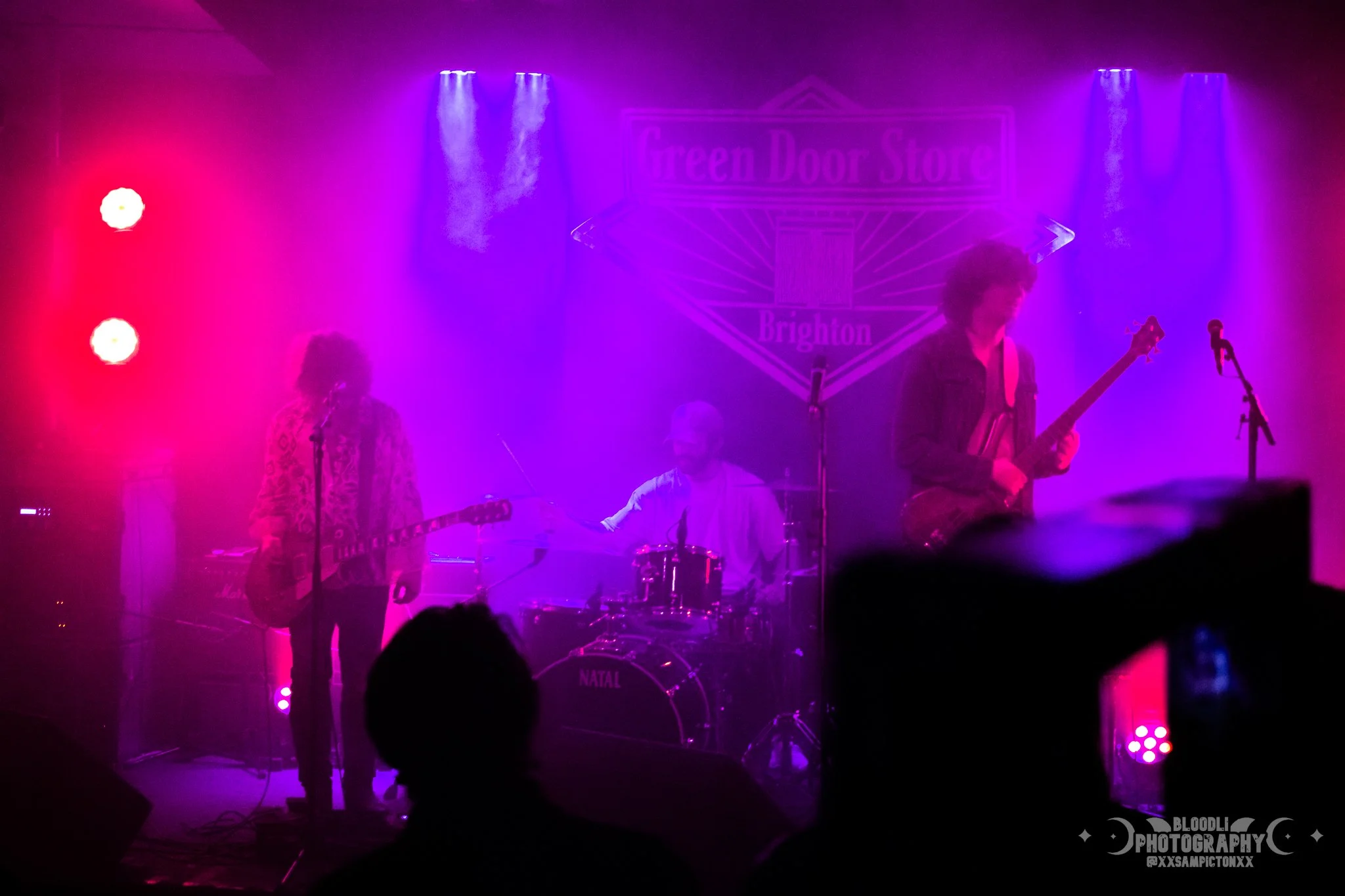 A band performing on stage with purple and pink lighting, featuring a guitarist, drummer, and bassist, with a sign that says 'Green Door Store Brighton' in the background.