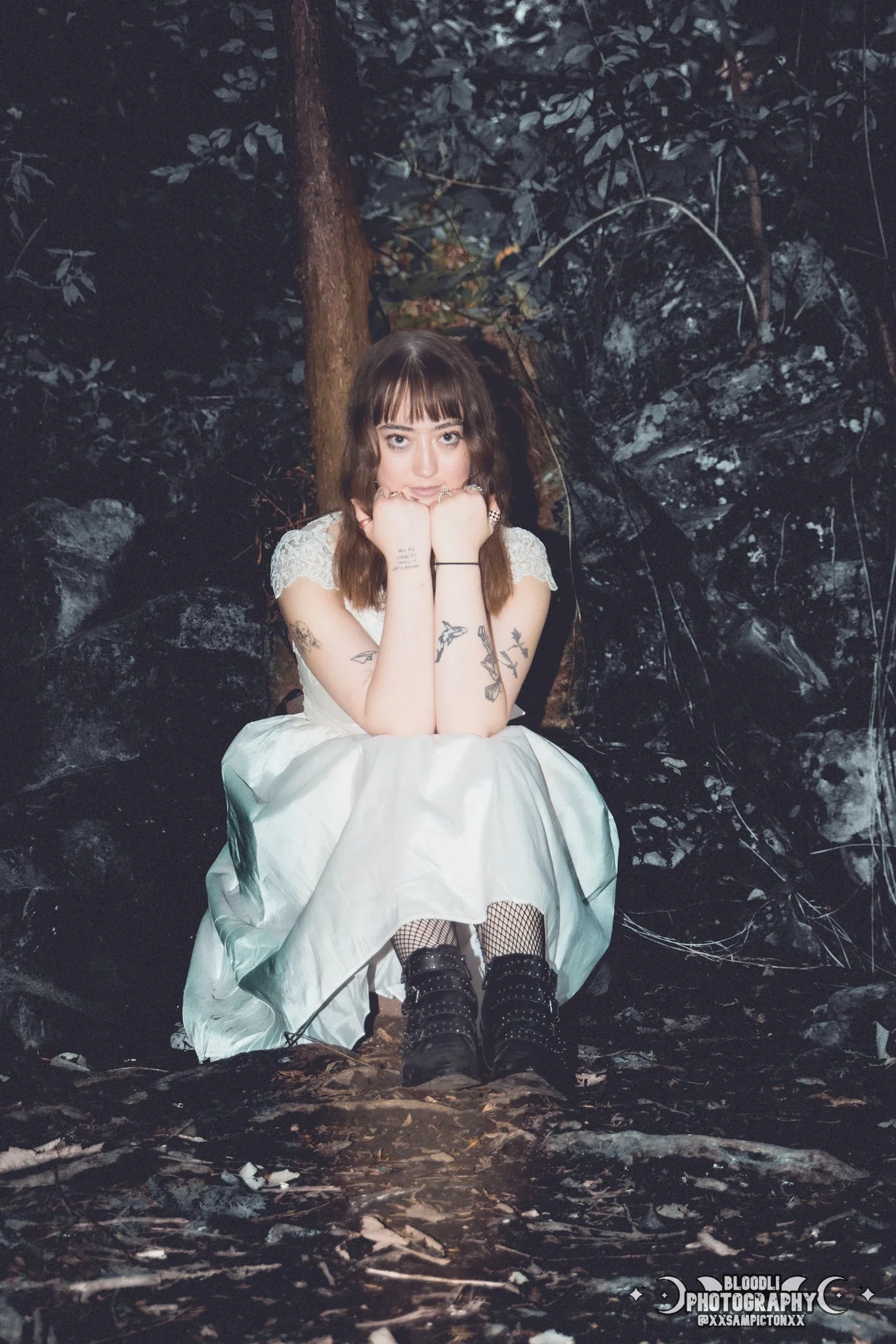 A young woman with tattoos on her arms and chest, wearing a white dress and black combat boots, sitting on the ground in a dark wooded outdoor setting, looking directly at the camera with her chin resting on her hands.