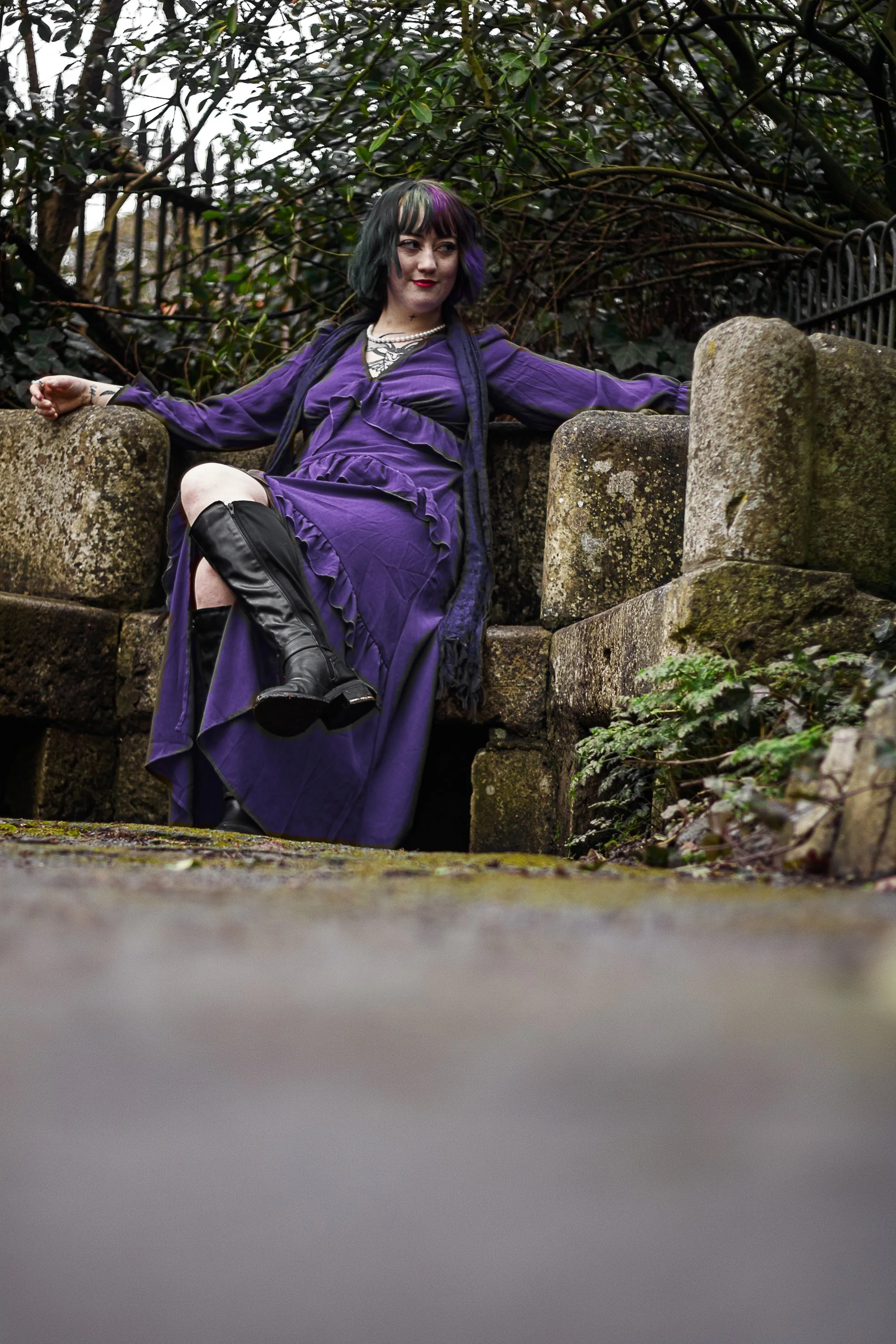 Woman in a purple dress and black boots sitting on a stone wall outdoors with greenery in the background.
