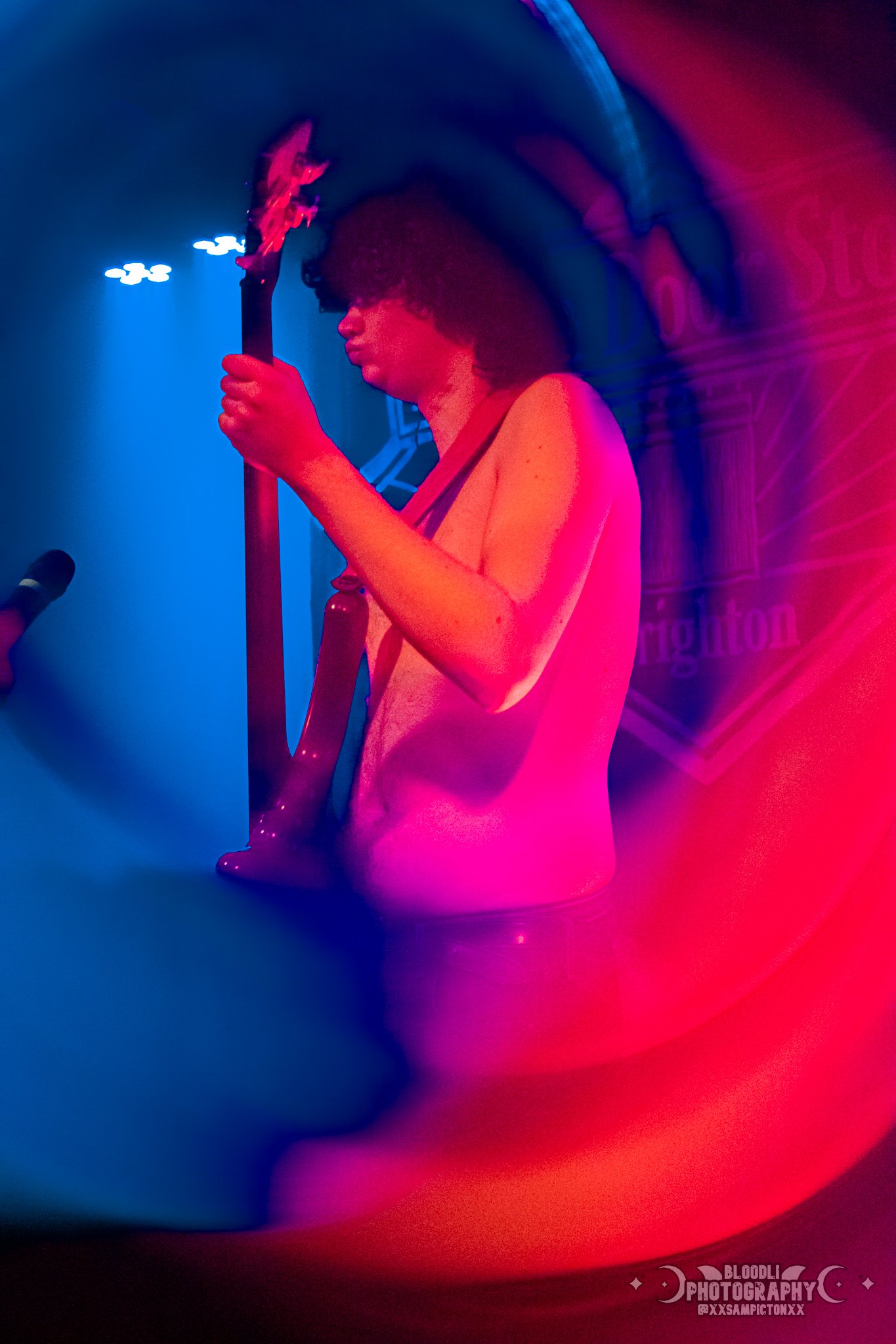 A person with curly hair playing an electric guitar on stage during a performance, illuminated with colorful pink and blue lighting.