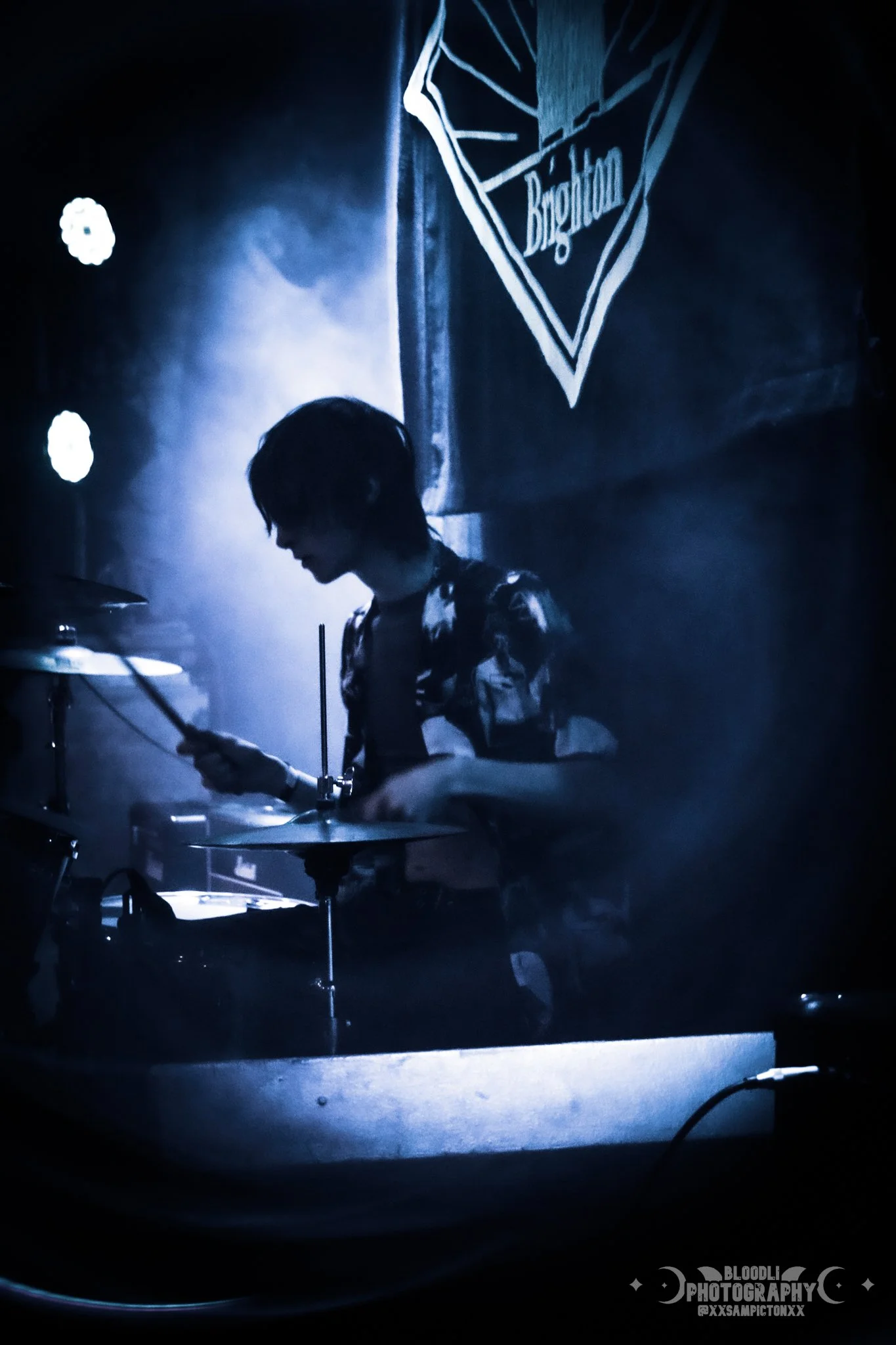 A person playing drums on stage, illuminated in blue light, with a Brighton banner in the background.