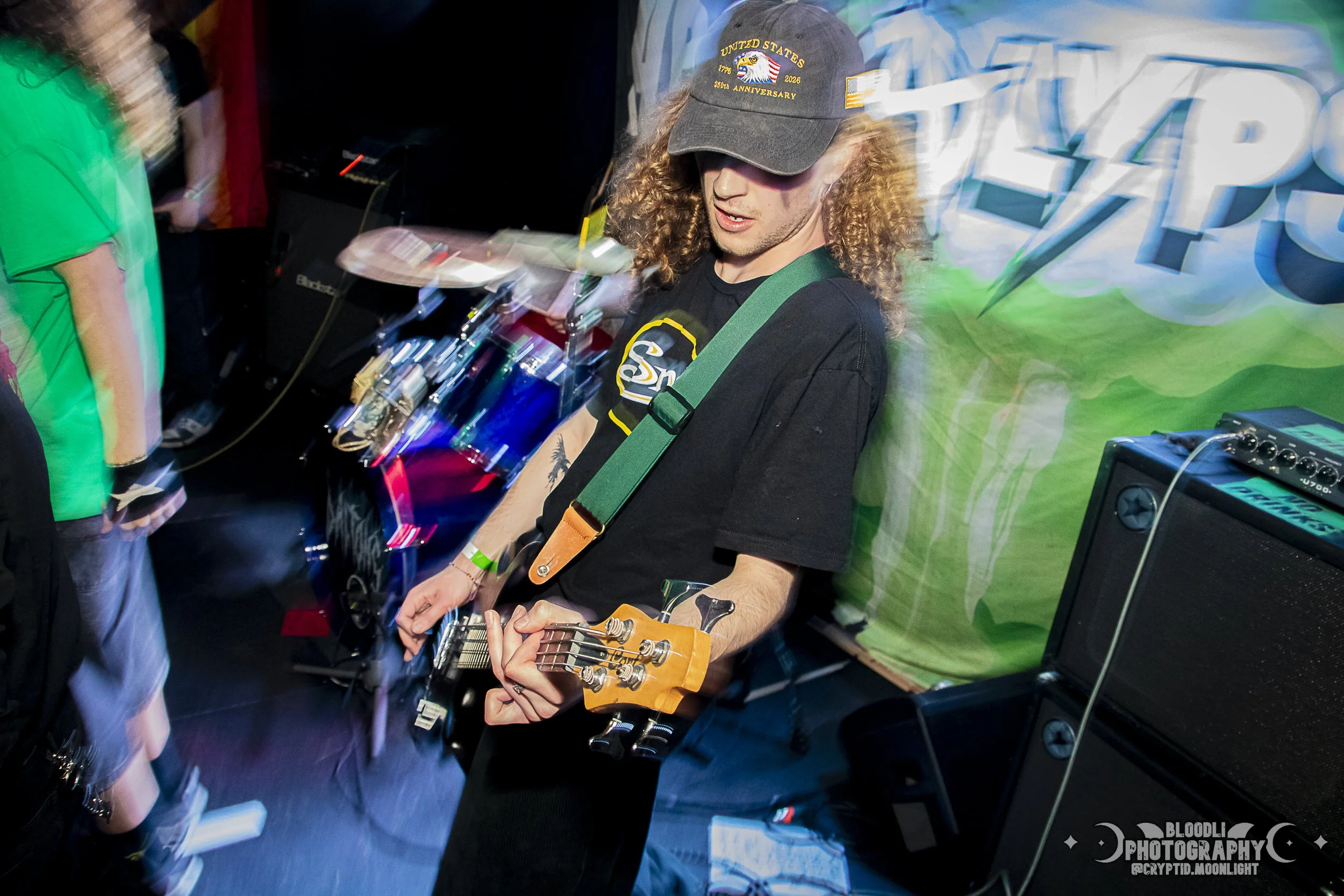 A drummer and a guitarist playing at a music event with a green backdrop and a banner. The guitarist, a young man with long curly hair, is wearing a baseball cap and a black t-shirt, focused on his guitar.