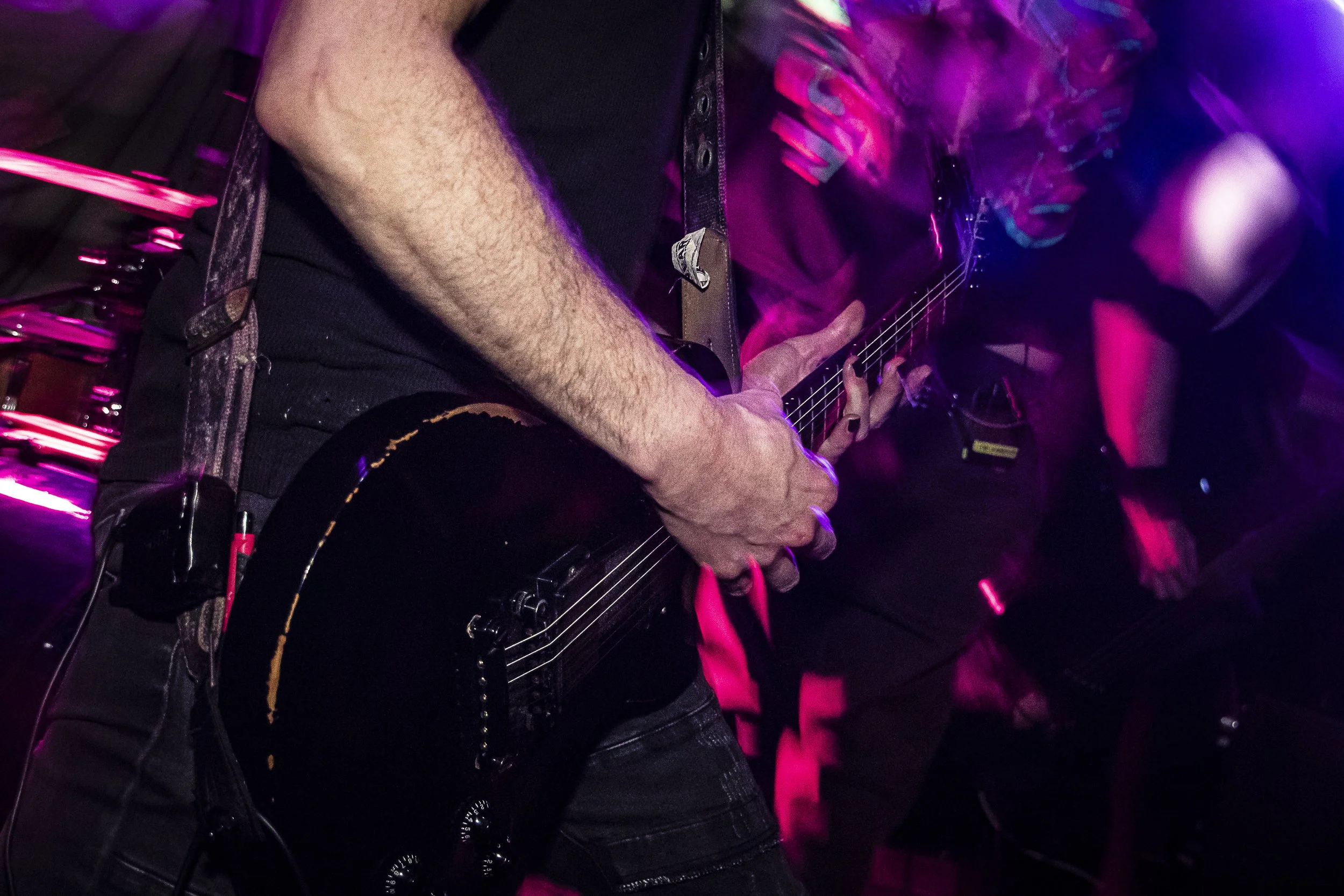 Close-up of a person playing an electric guitar at a music venue with vibrant pink and purple lighting.