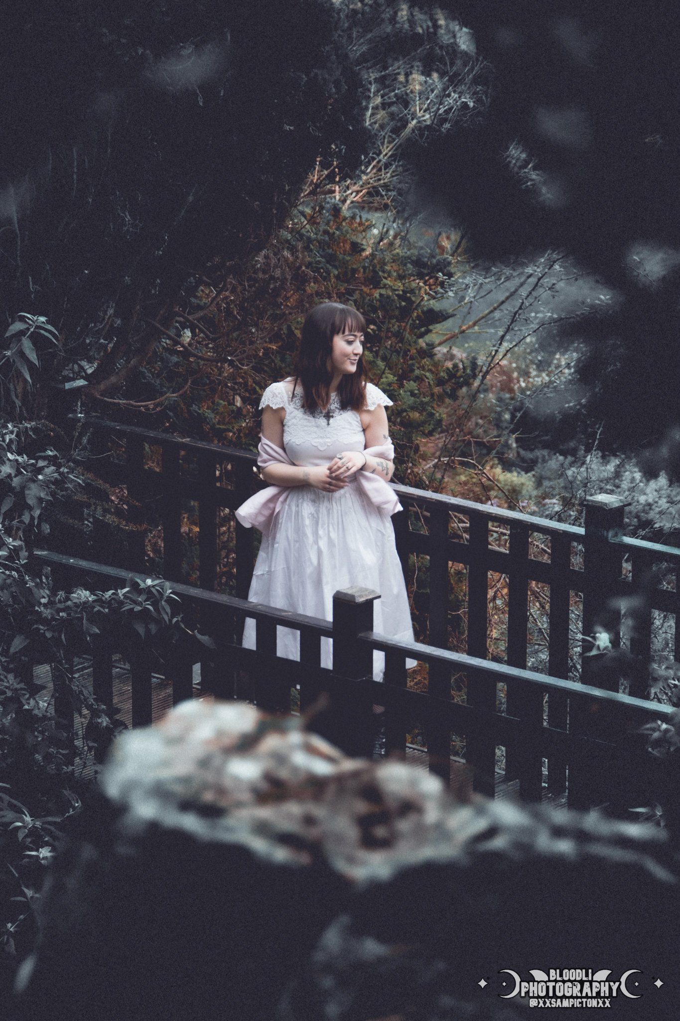 A young woman in a white dress standing on a wooden bridge in a forest, looking to the side with a peaceful expression.