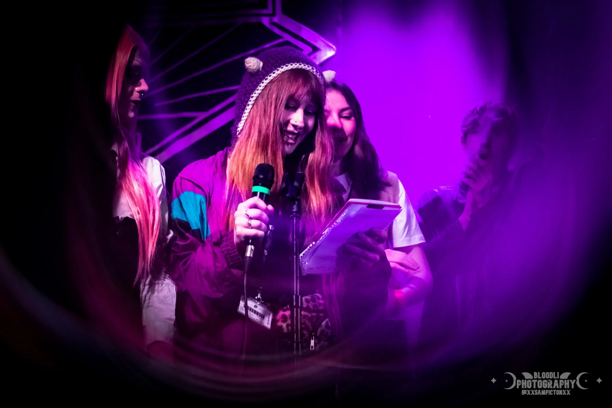 Four young women on stage, one with a microphone, reading from a notebook, under purple lighting.