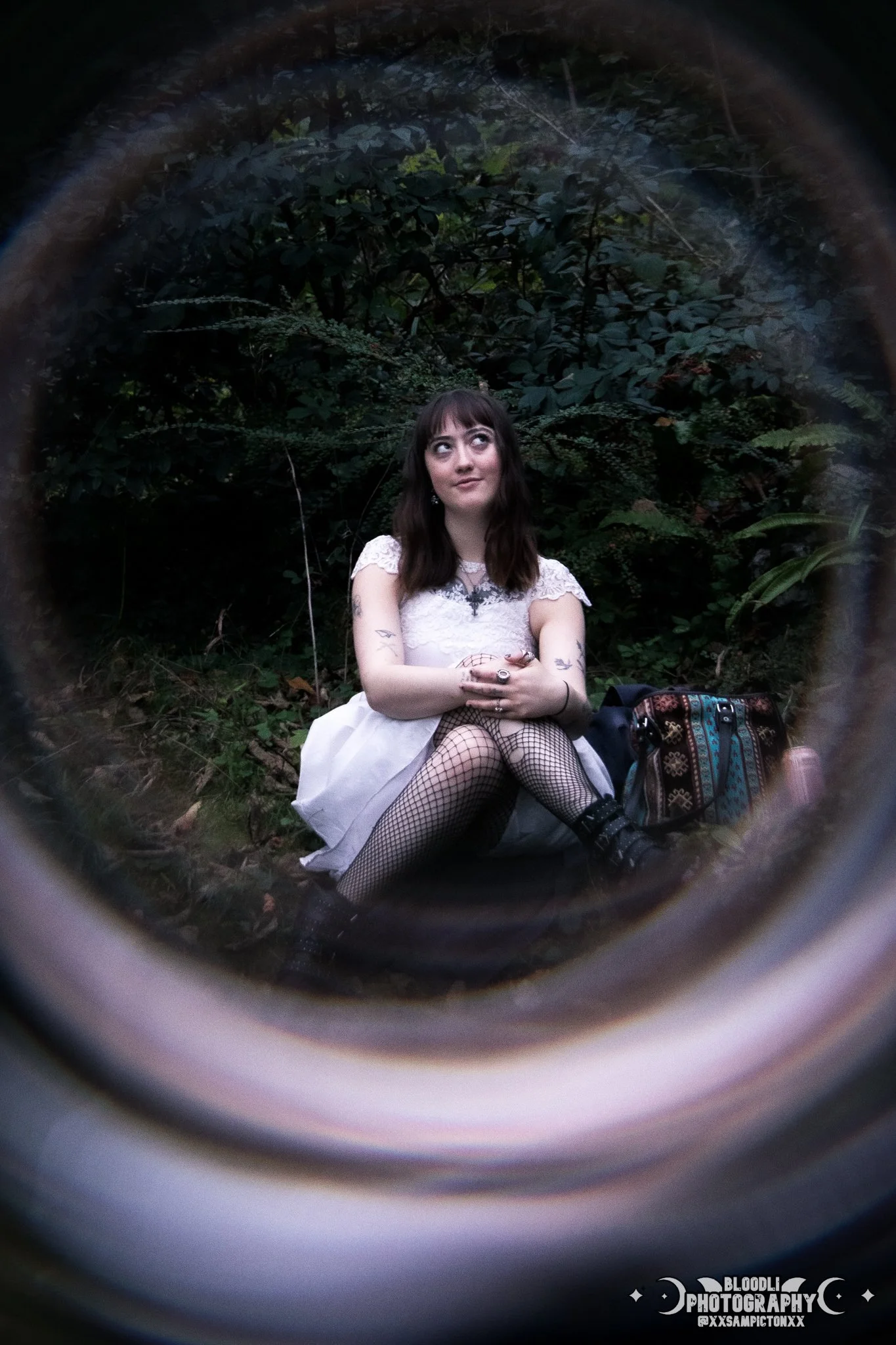 A young woman with dark hair, tattoos, fishnet stockings, and goth-style jewelry sitting outdoors in front of lush green foliage, with a backpack beside her.