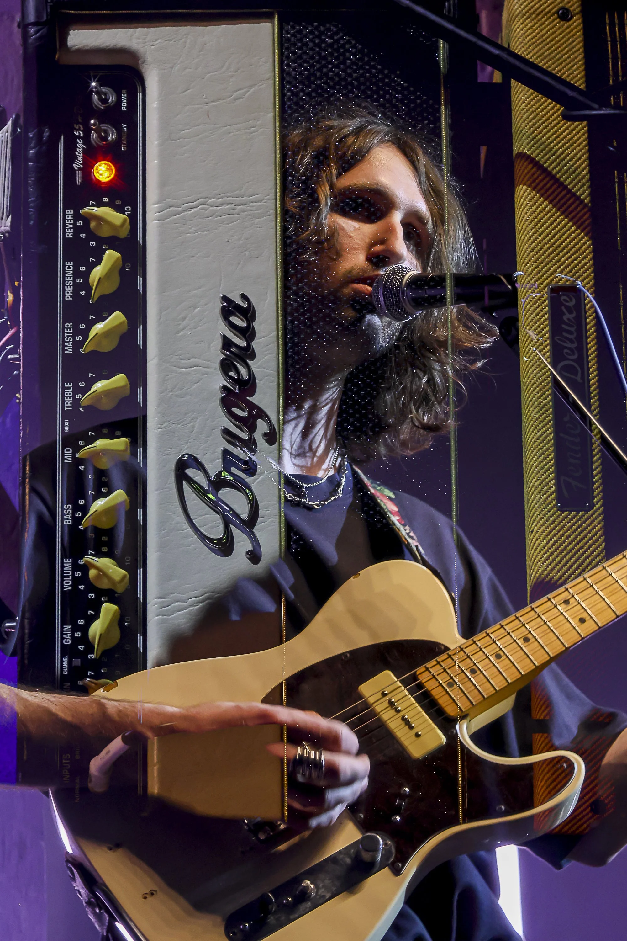 A musician playing an acoustic guitar while singing into a microphone. The image is reflected in a mirror that also shows a vintage bass amplifier with yellow control knobs and a Fender brand logo.