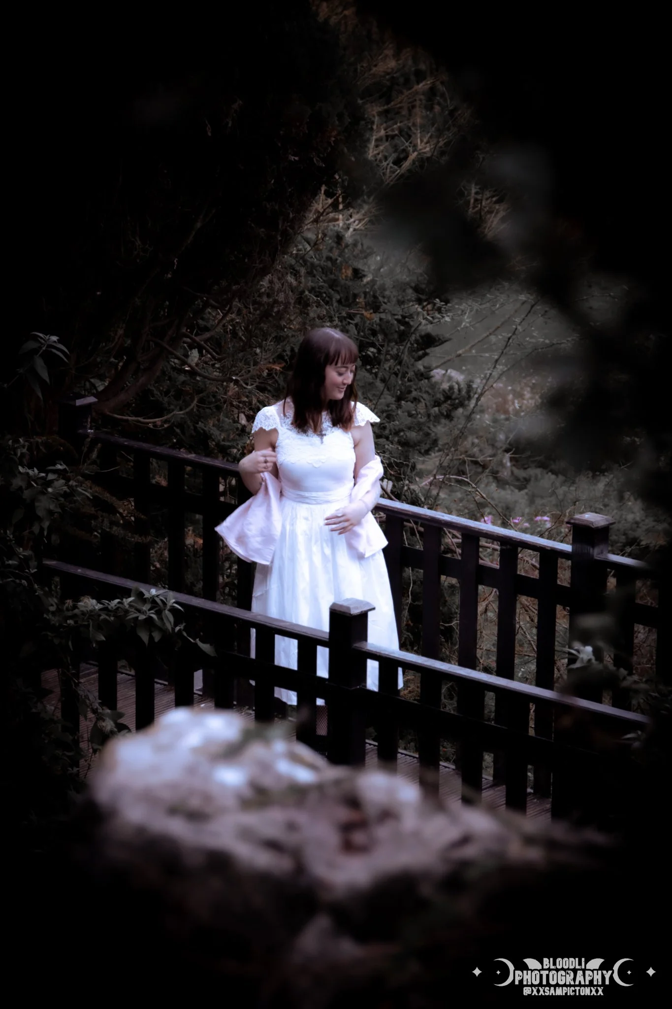 A woman in a white dress standing on a wooden bridge in a forested area.