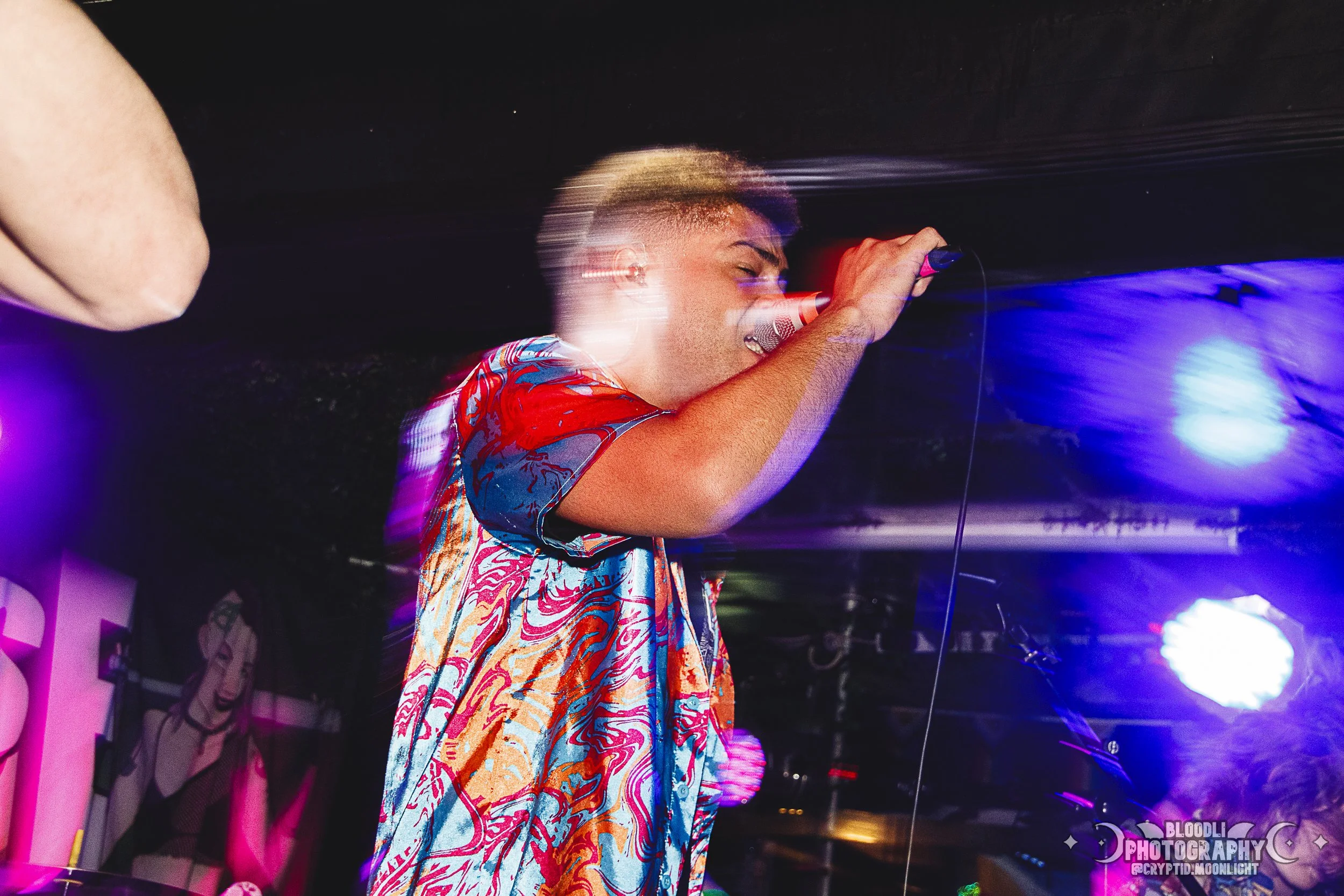 Male singer performing with a microphone in a vibrant, colorful setting, wearing a patterned shirt, with stage lights creating a dynamic atmosphere.