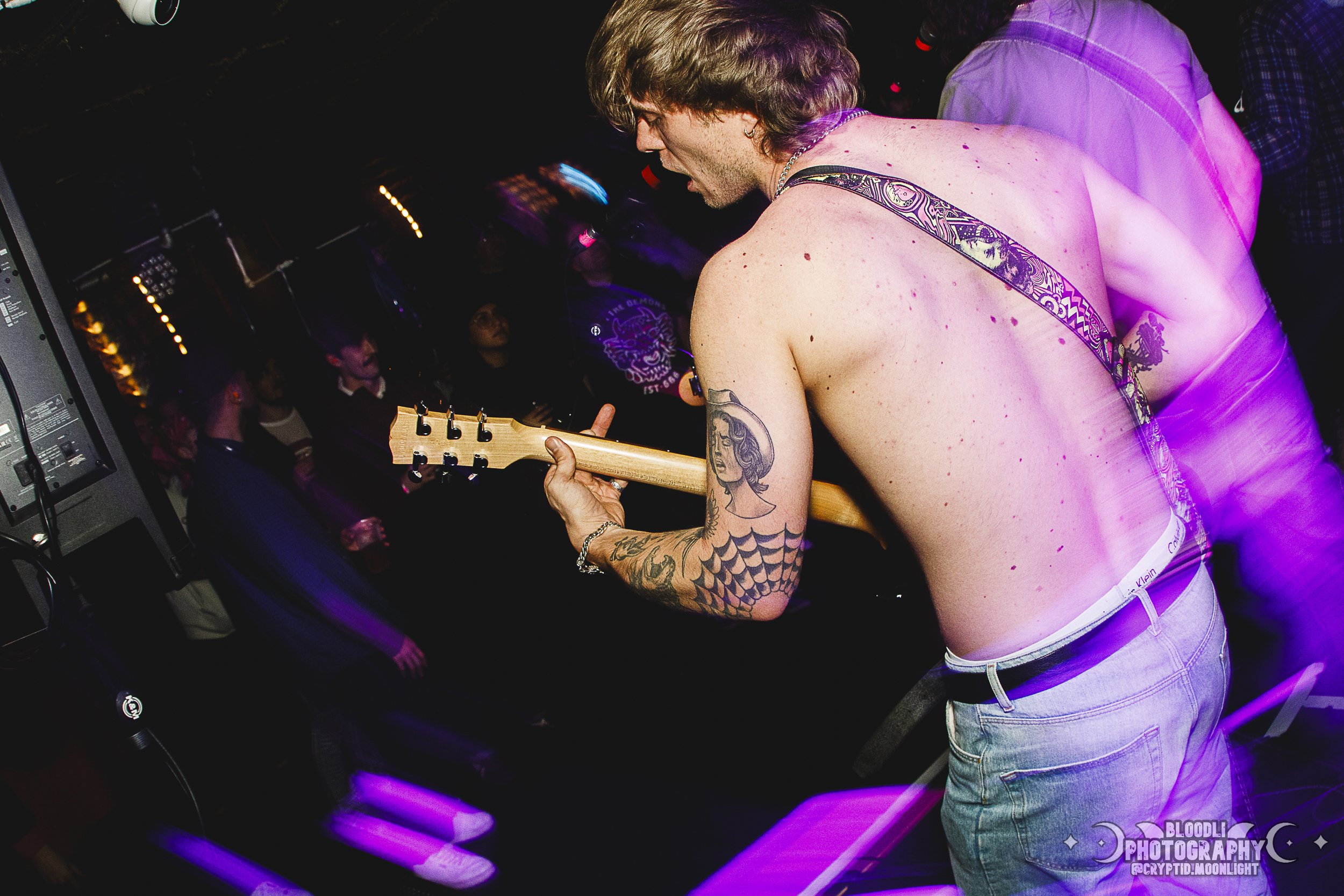 A shirtless man playing an electric guitar at a nightclub, with tattoos on his arms, surrounded by a crowd of people.