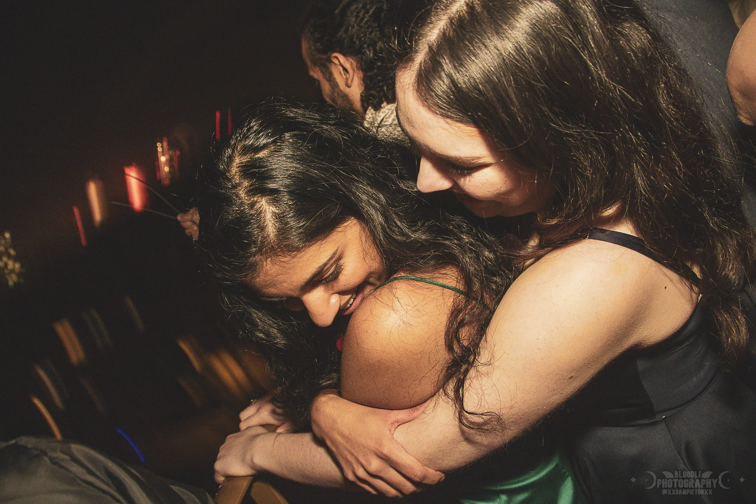 Two women embrace each other and smile in a close, intimate moment at a party or nightclub with dim lighting and blurred background.
