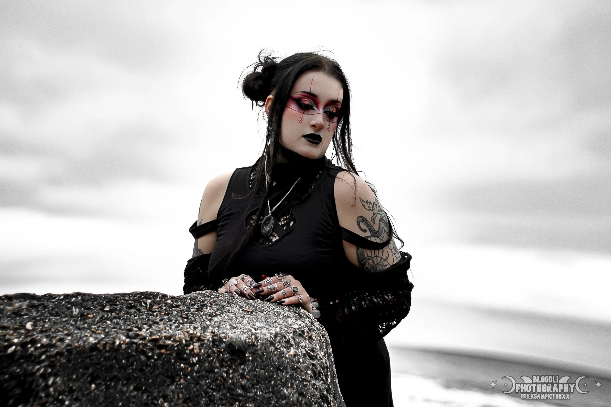 A woman with gothic style makeup and black hair styled in buns, wearing a black sleeveless top with chain accessories and multiple rings, leaning on a textured rock with tattoos on her arm, against a cloudy sky background.
