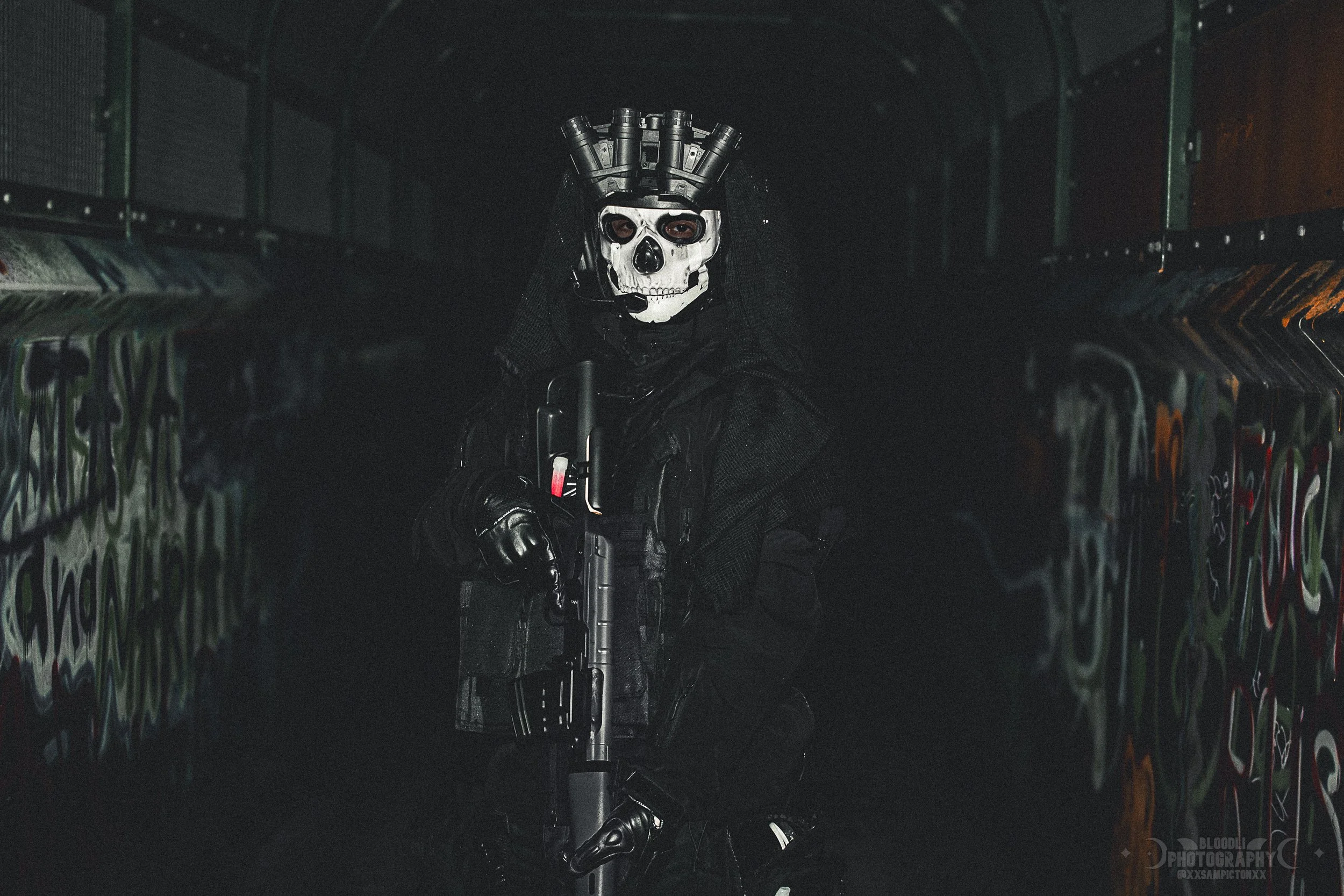 Person in tactical black gear kneeling in a graffiti-covered underpass, wearing a skull mask with what appears to be grenade shells on the head, holding a firearm.