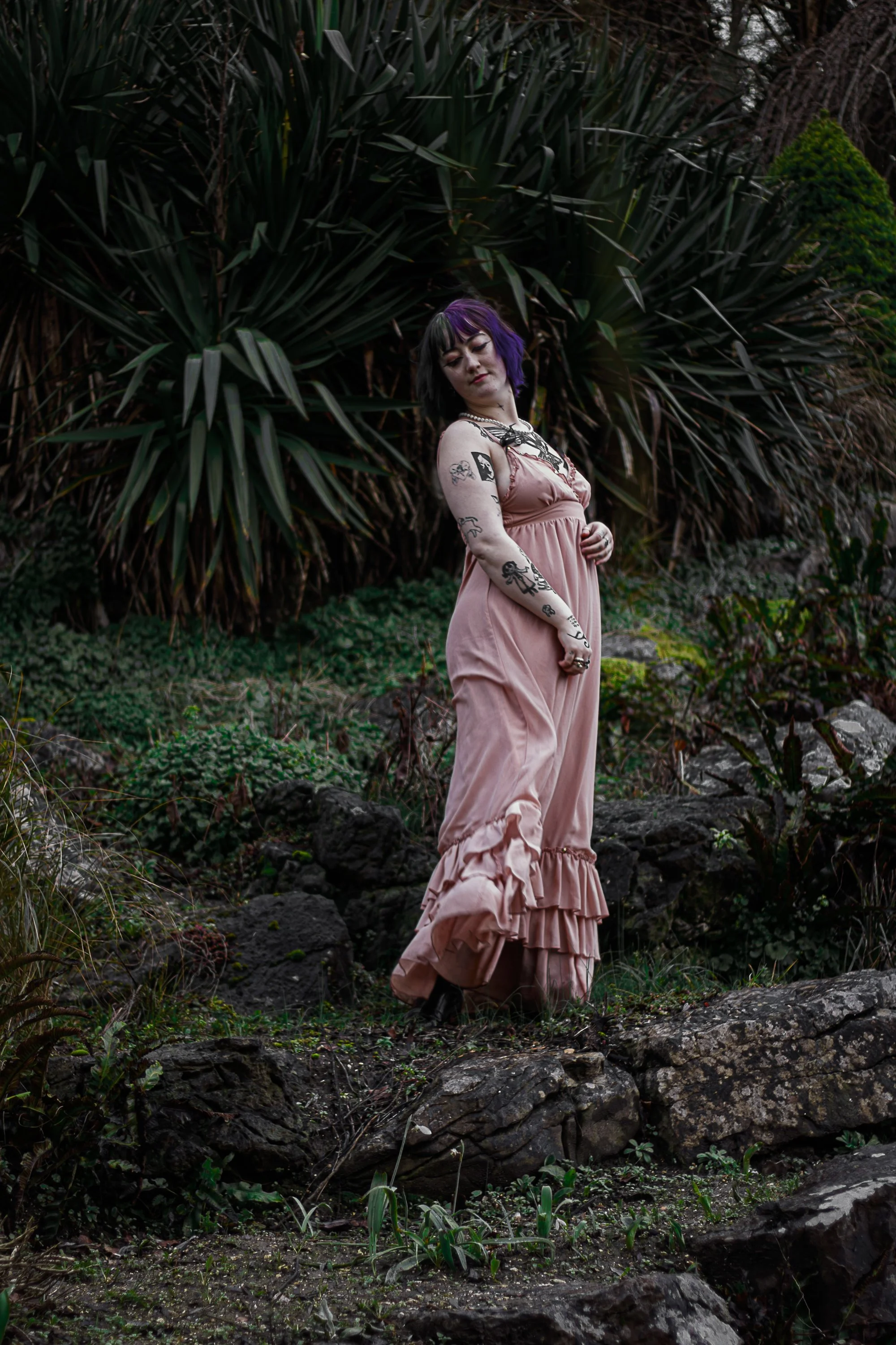 A woman with tattoos and purple and black hair in a pink ruffled dress standing on rocks amidst green plants and trees in a natural outdoor setting.