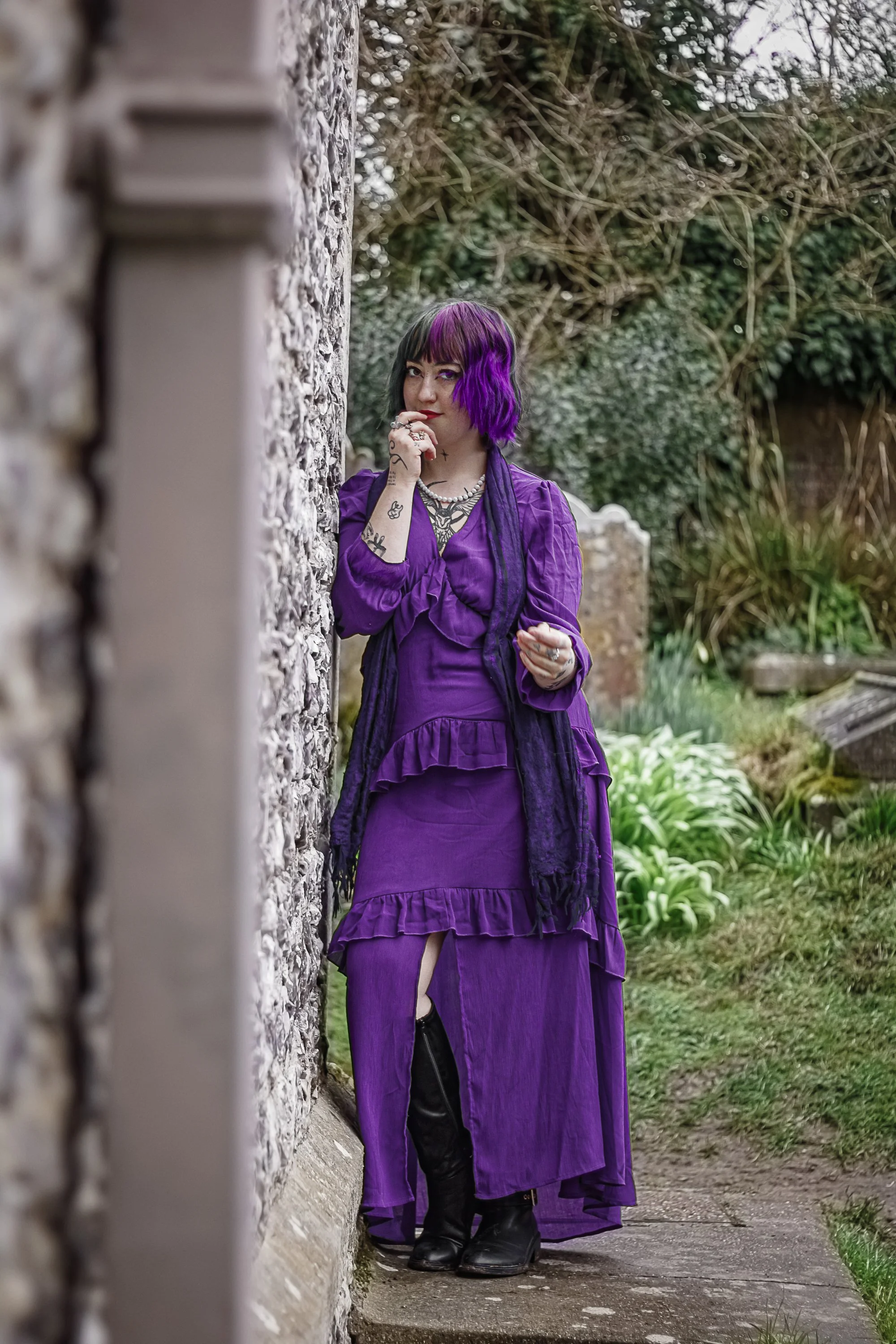 A woman with purple and black hair, tattoos, and wearing a long purple dress with ruffles, black boots, and layered jewelry, standing next to a stone wall and holding her chin thoughtfully in a garden setting.