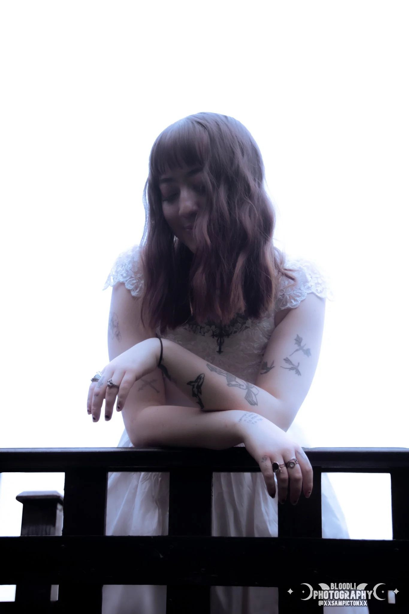 A woman with shoulder-length wavy brown hair, tattoos on her arms, wearing a white dress with lace on the sleeves, leaning on a dark wooden railing with a bright white background.