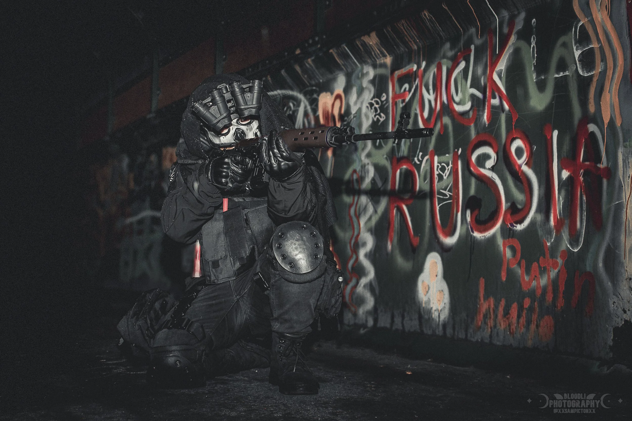 A person dressed in tactical gear, wearing a mask with several camera lenses, kneeling and aiming a rifle at a graffiti-covered wall with a spray-painted message that includes profanity, in a dimly lit urban setting.