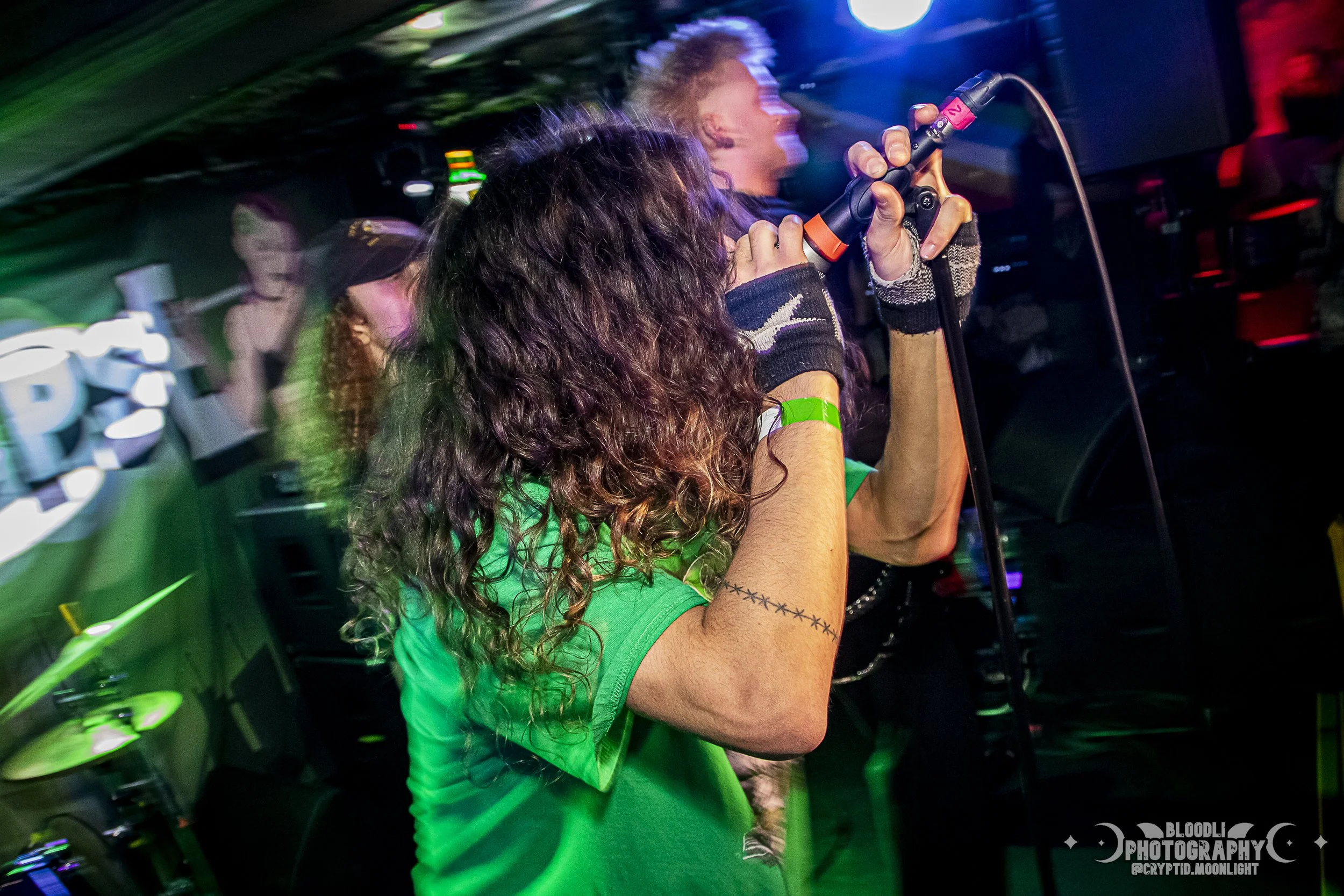 Person with curly hair singing into a microphone on stage at a concert or club, with colorful lights and other people in the background.