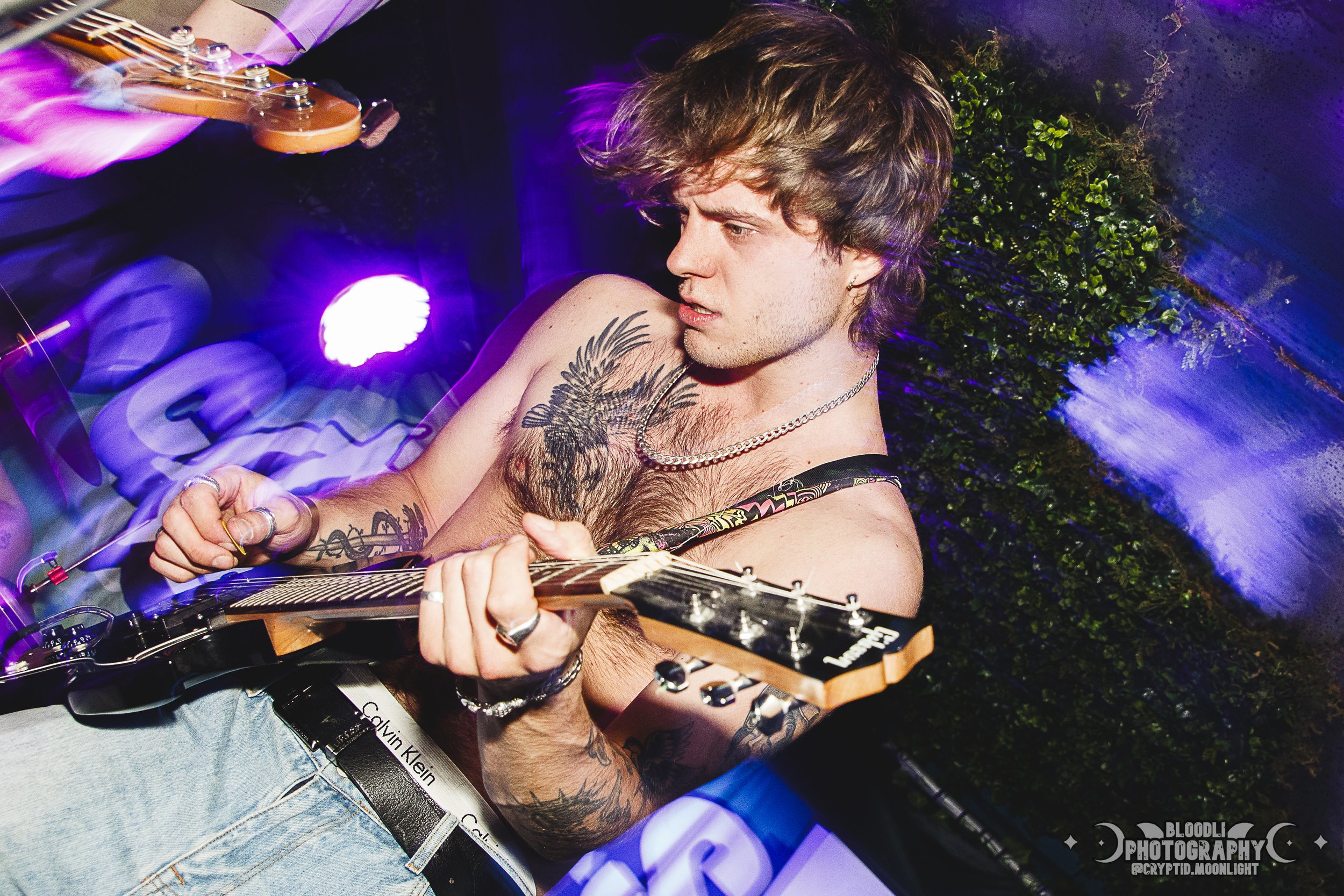 A shirtless young man with tattoos, wearing a chain necklace and Calvin Klein underwear, is playing an electric guitar in a vibrant, colorful setting with purple and blue lighting.