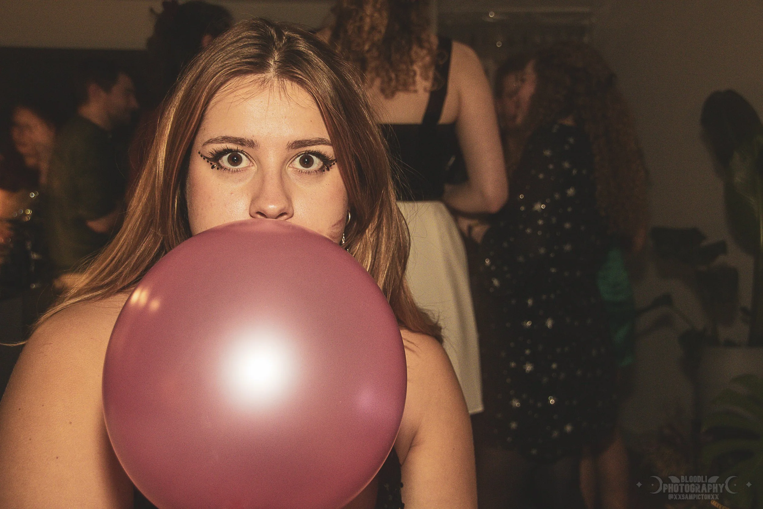 A young woman with makeup and long hair holding a pink balloon in front of her lips at a party, with other people in the background.