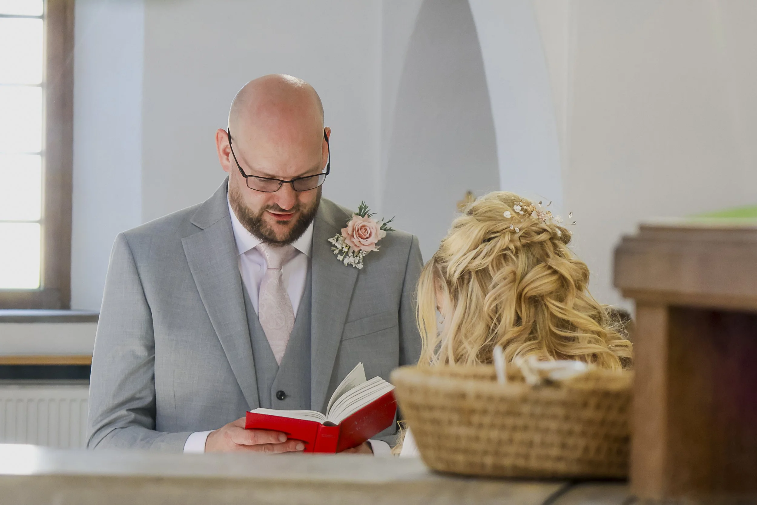 A man in a gray suit and glasses is reading a red book, with a woman with blond curly hair and floral hair accessories beside him, in an indoor setting with a window and staircase in the background.