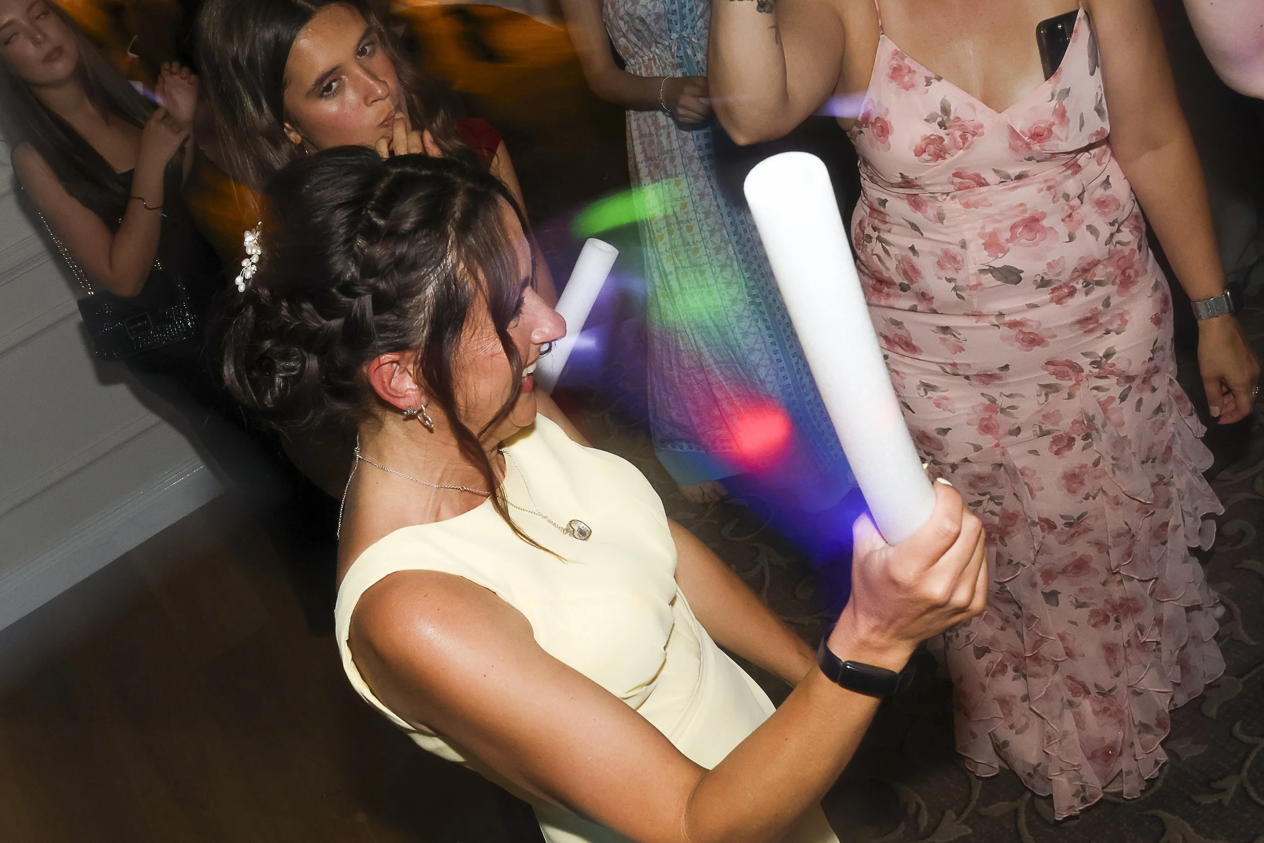 A woman wearing a yellow dress with curly hair styled with a braid and floral hair accessory, holding a glowing party tube while smiling, at a social event with other women in the background.
