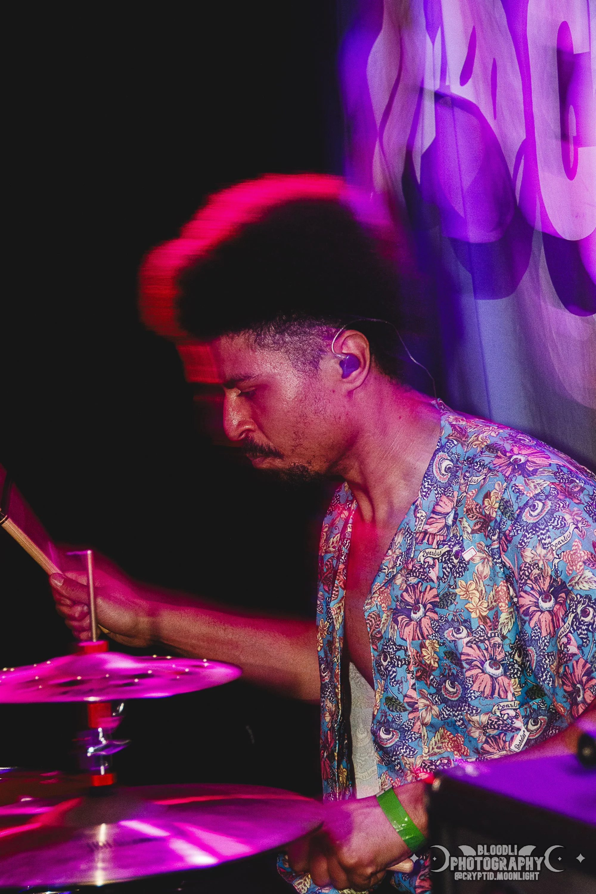 A man with a high-top hairstyle plays drums at a concert, wearing a colorful floral shirt and a green wristband, with purple and pink lighting.