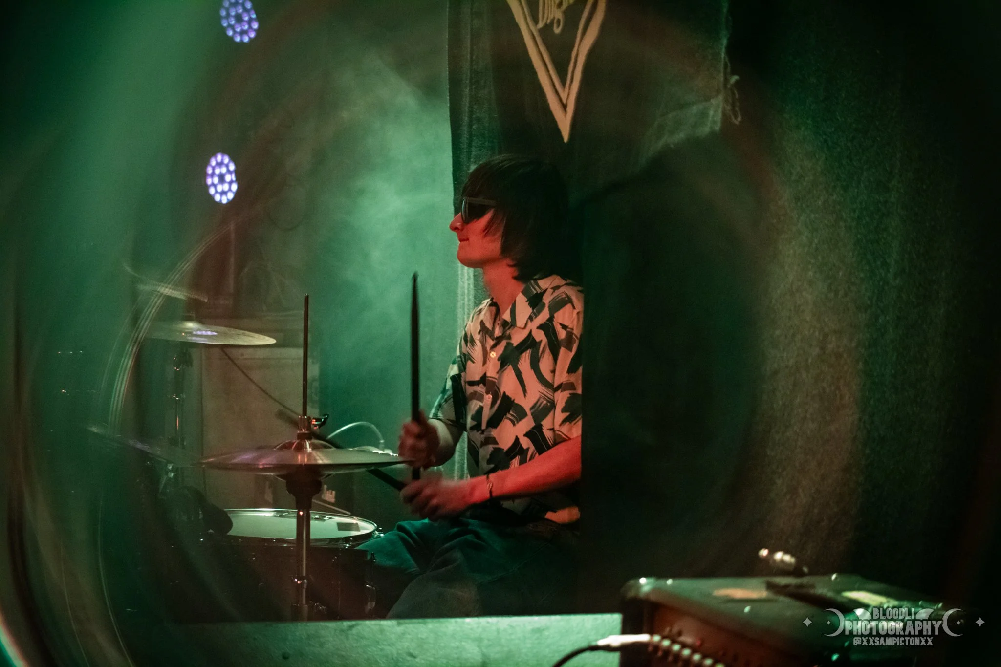Side view of a person with dark hair wearing sunglasses and a patterned shirt, sitting with a drumstick in hand next to a drum set, illuminated by green and purple stage lights.