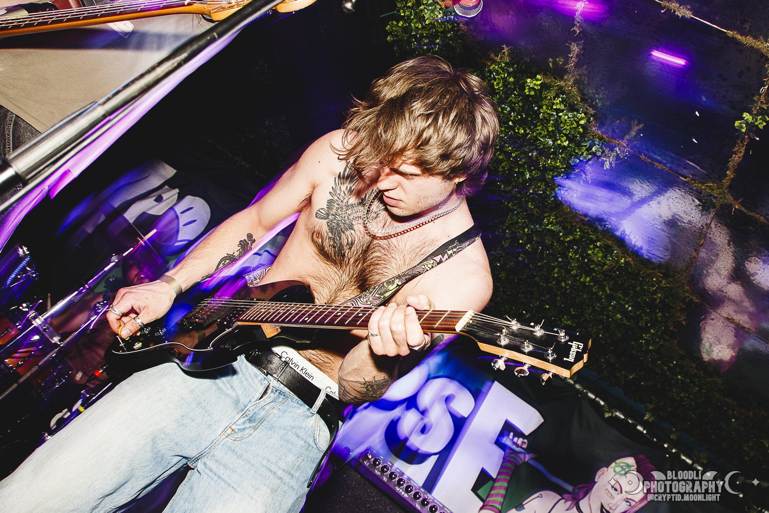 A shirtless young man with shoulder-length hair playing an electric guitar on stage, with tattoos visible on his chest and arm. The stage has colorful lighting and a backdrop with greenery and signage.