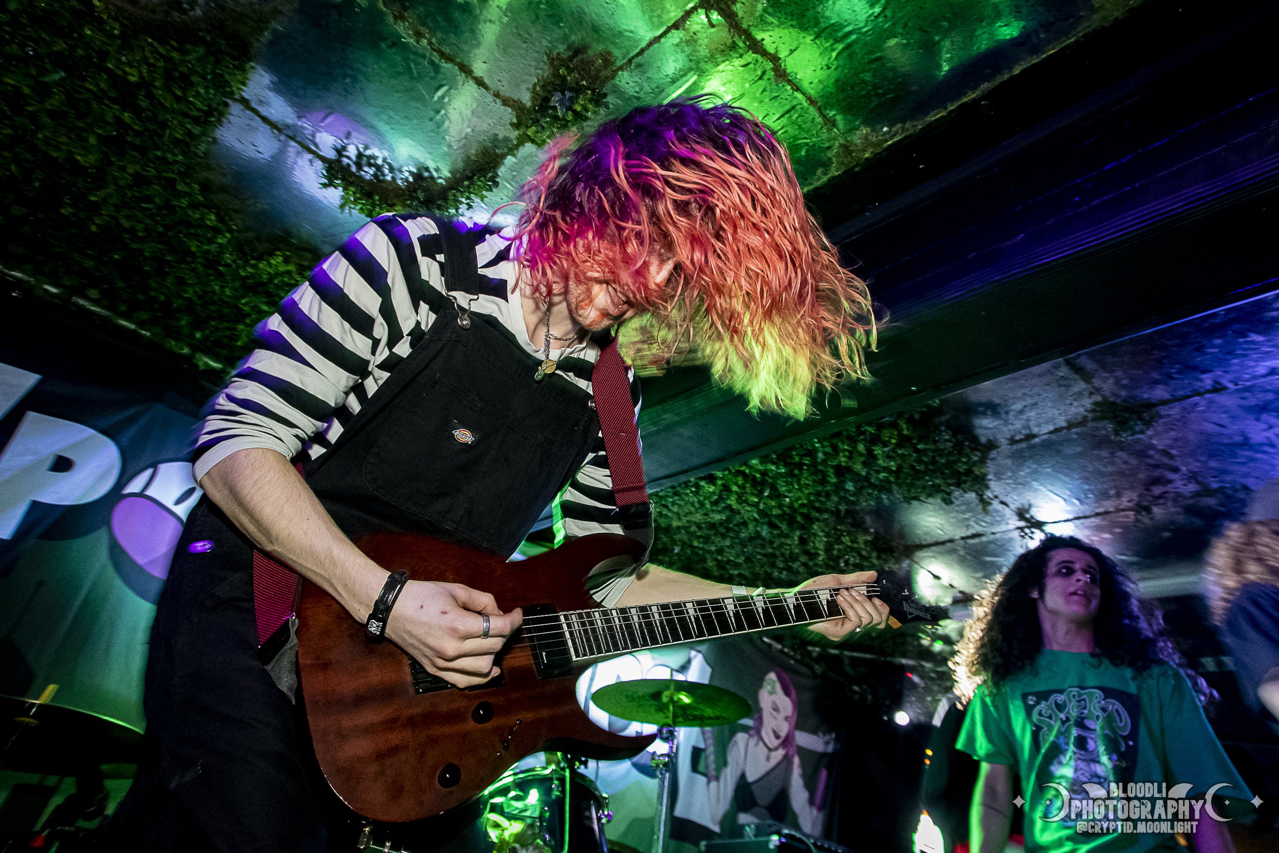 A musician with curly, brightly colored hair playing an electric guitar during a live performance, with a woman with curly hair and a colorful shirt near a drum set in the background, under colorful stage lights.