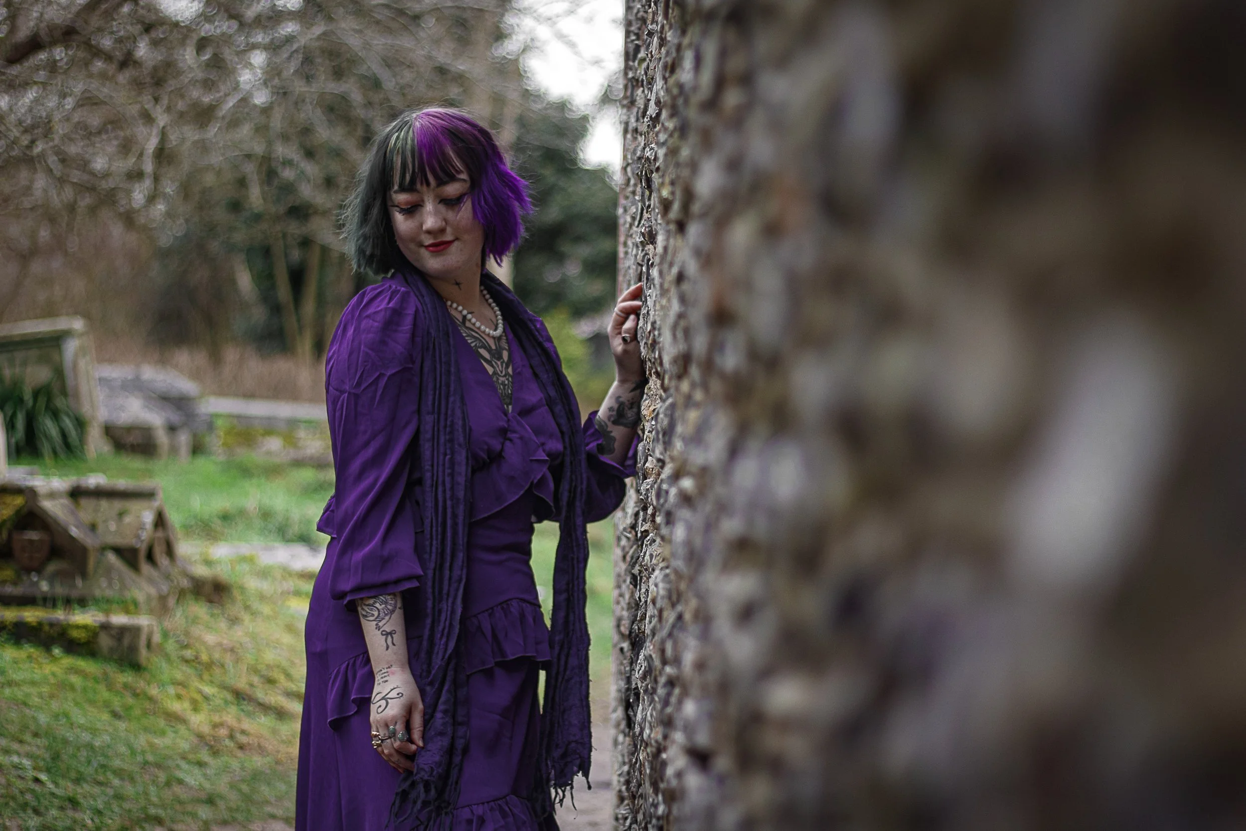 A young woman with black and purple hair, dressed in a purple dress, stands outdoors next to a stone wall, looking down and smiling.